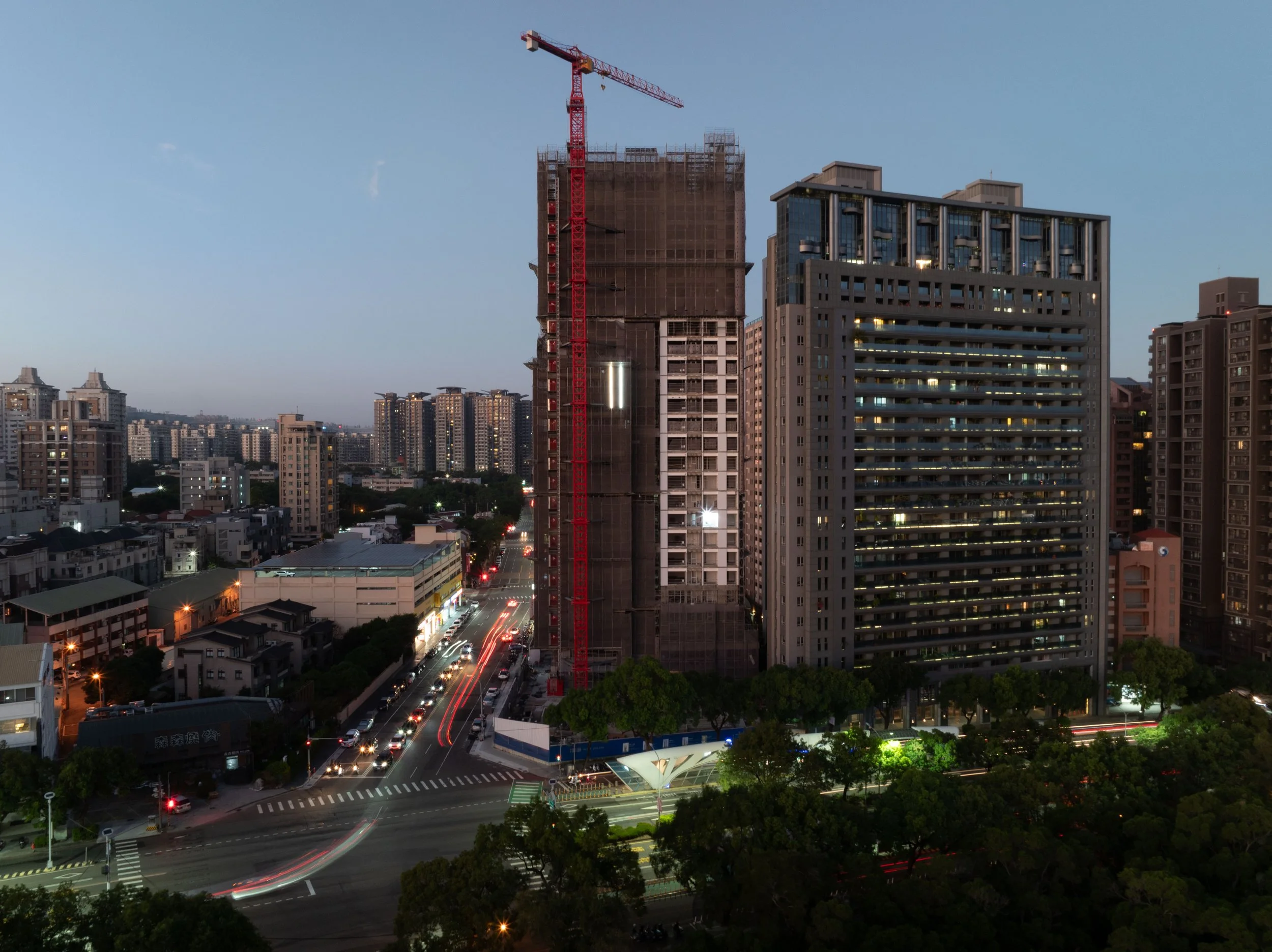 Cityscape at dusk featuring high-rise buildings under construction with a red crane, busy busy intersection with light trails from vehicles, and surrounded by trees and other buildings.
