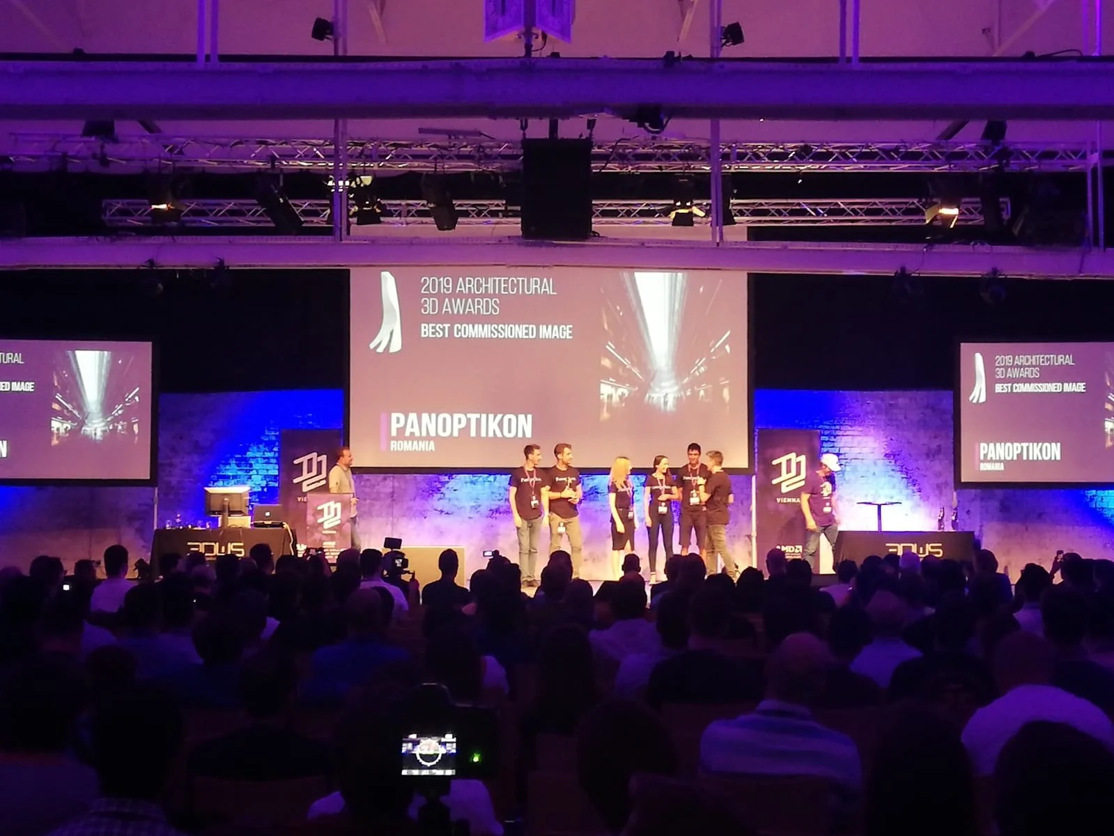 People on stage at the 2019 Architectural 3D Awards ceremony, receiving the Best Commissioned Image award, with a large audience watching.