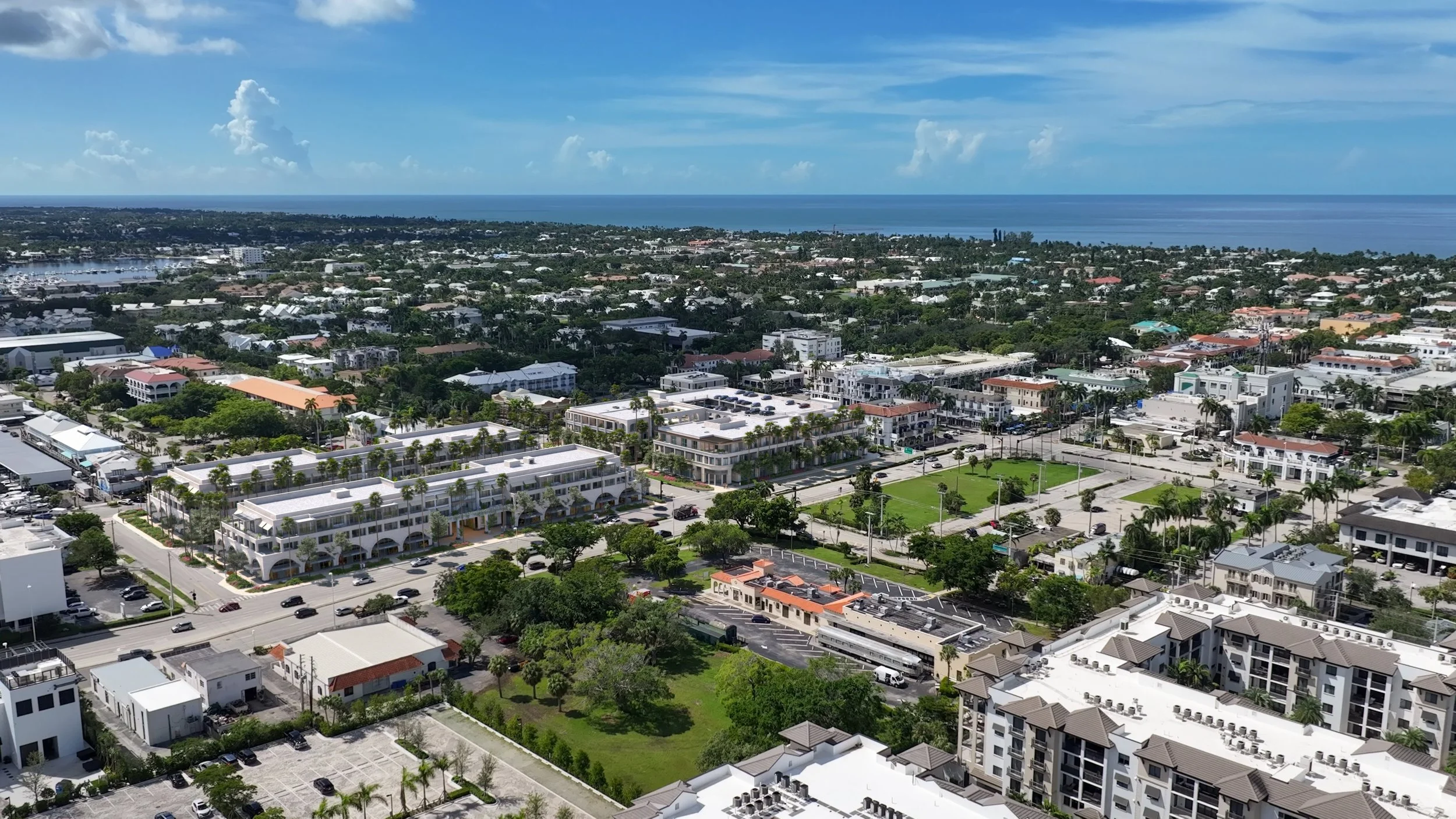 Aerial view of a coastal city with city buildings, green parks, and the ocean in the background under a partly cloudy sky.