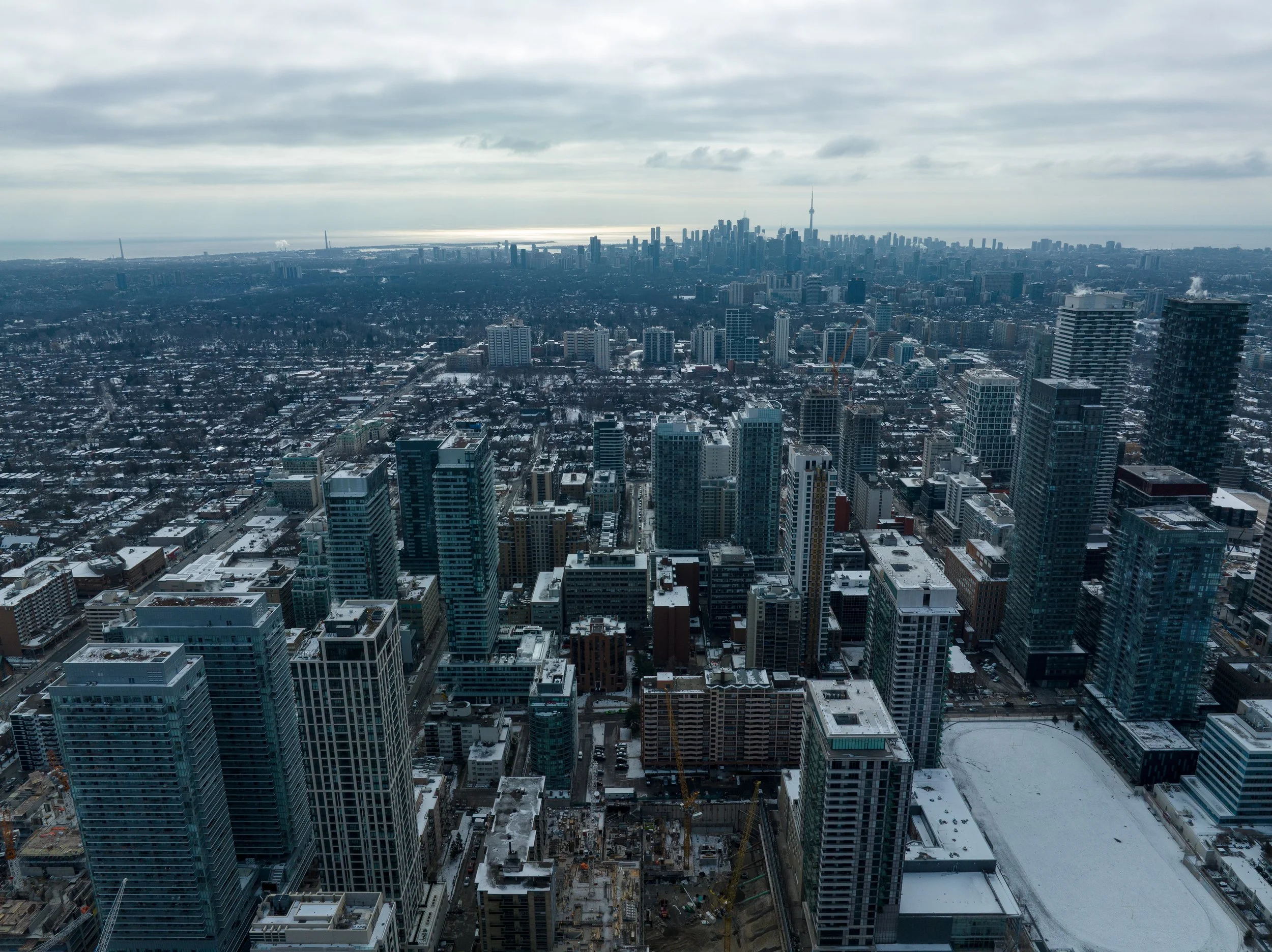 Aerial view of a city skyline in winter with tall modern skyscrapers, some under construction, snow-covered rooftops, and a cloudy sky.