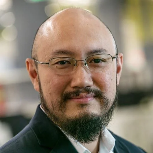 Close-up portrait of a man with glasses, a beard, and a bald head, wearing a suit jacket and white shirt, in an indoor setting.