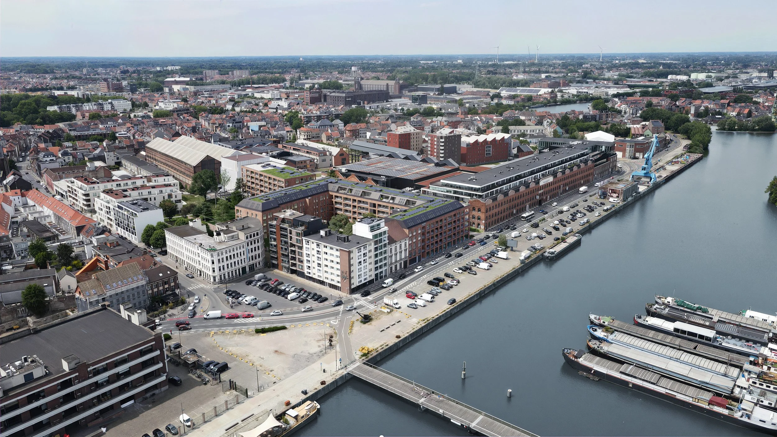 Aerial view of a city waterfront with boats docked, parking lots, modern and historic buildings, and a crane along the water's edge.