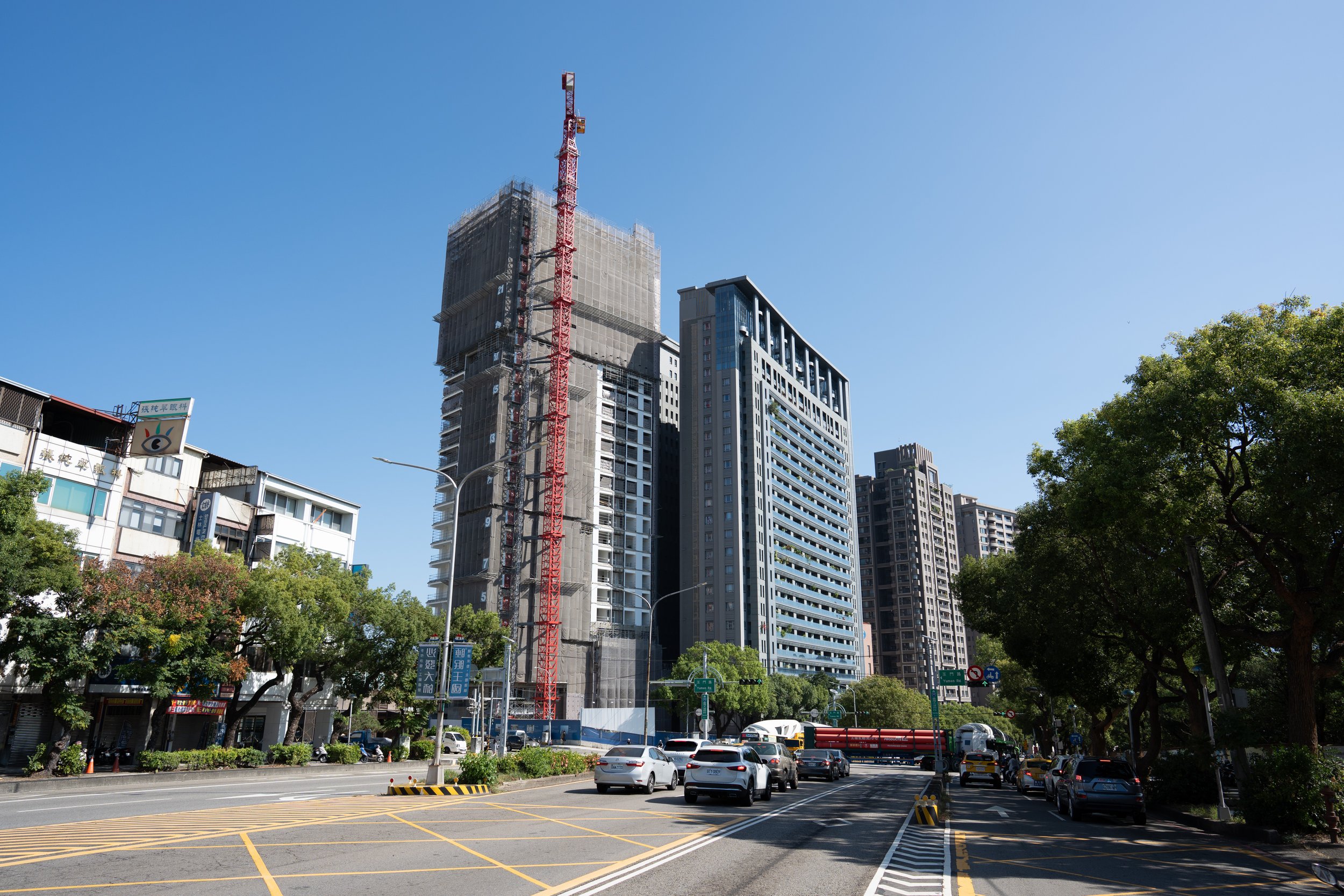 Urban street scene with construction site in the background, several completed buildings, cars parked along the road, and trees lining the sidewalk under a clear blue sky.