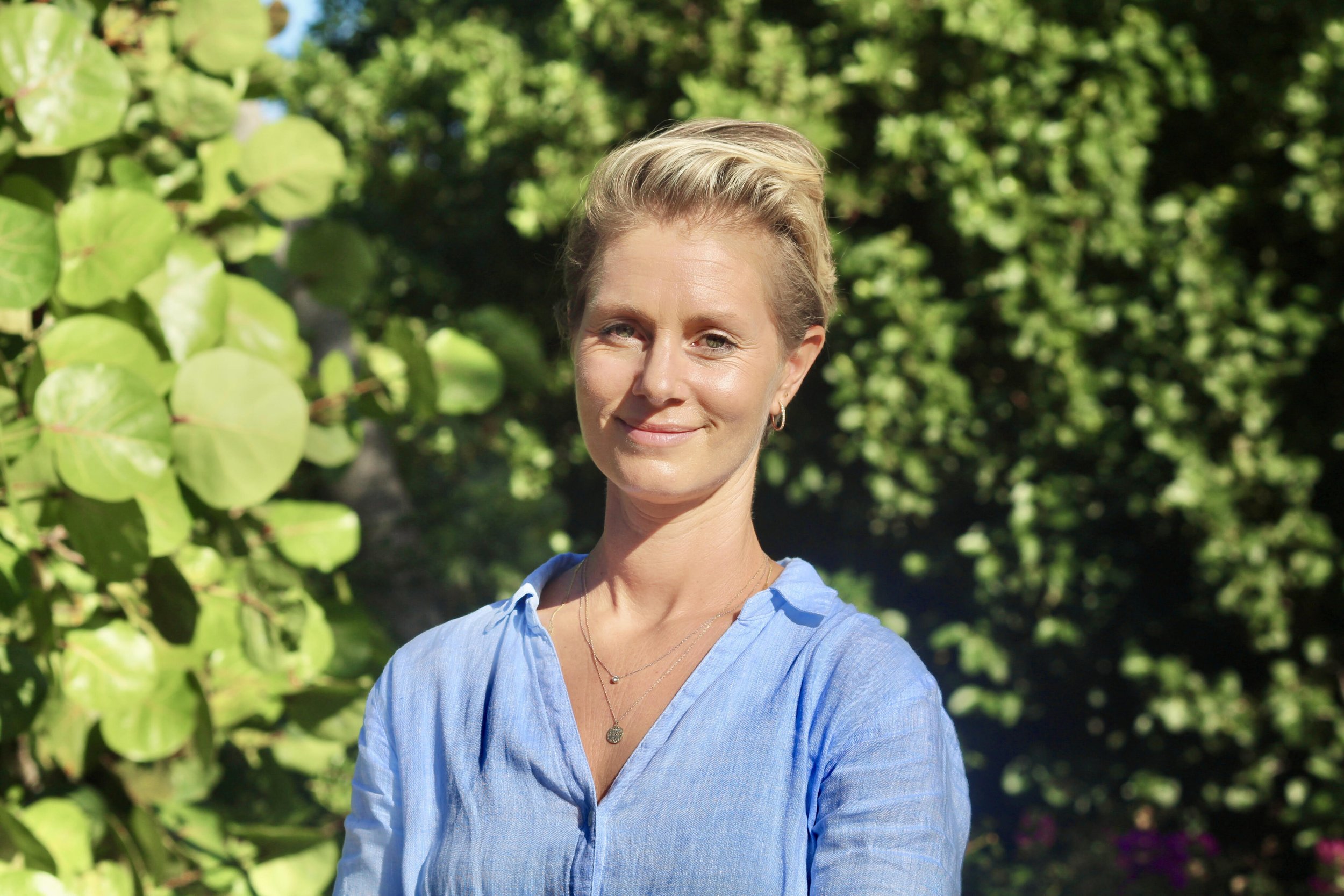 A woman with short blonde hair wearing a light blue shirt standing outdoors in front of green trees and plants.
