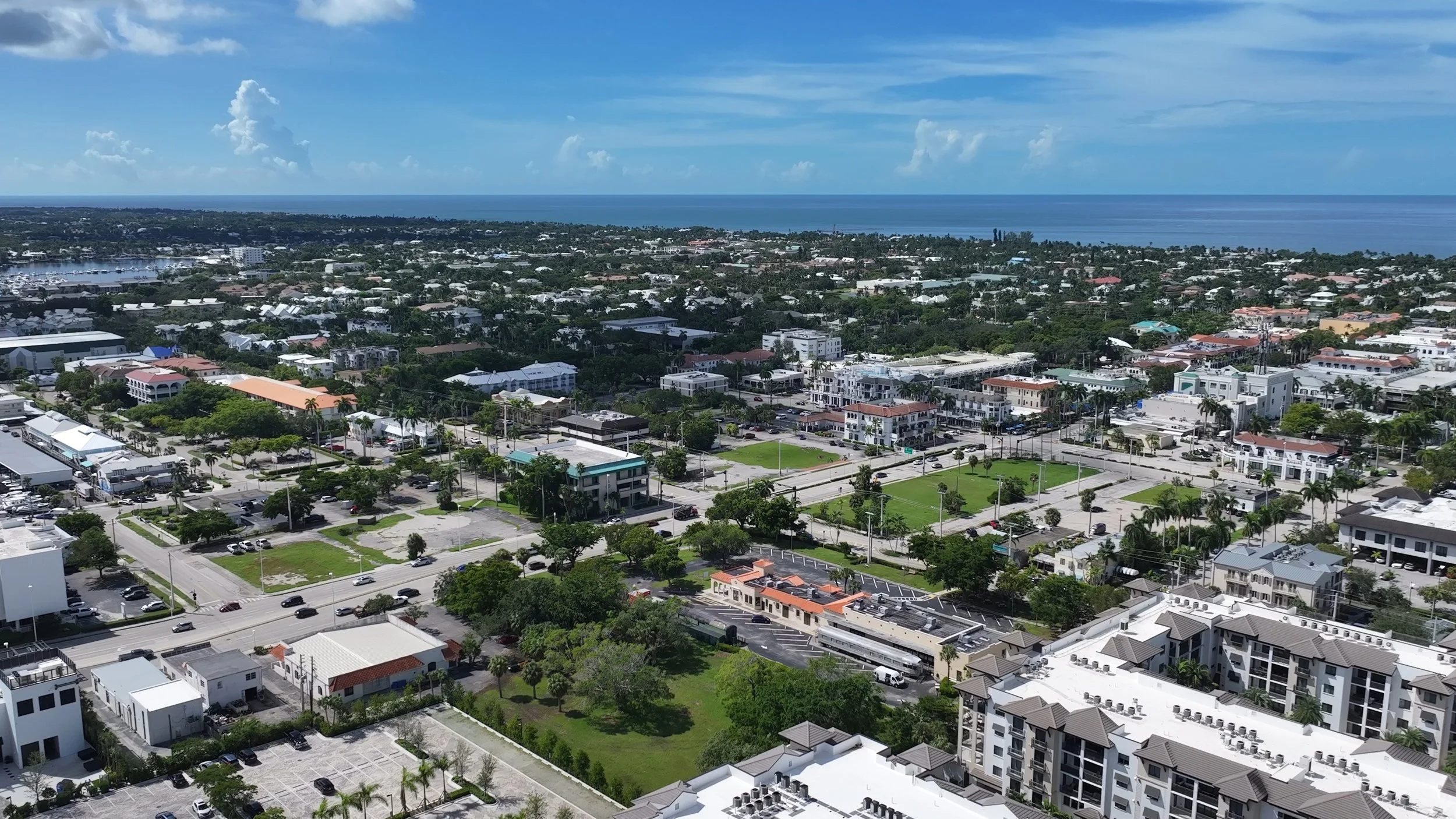 Aerial view of a cityscape near the coast with buildings, green parks, and a view of the ocean in the background.