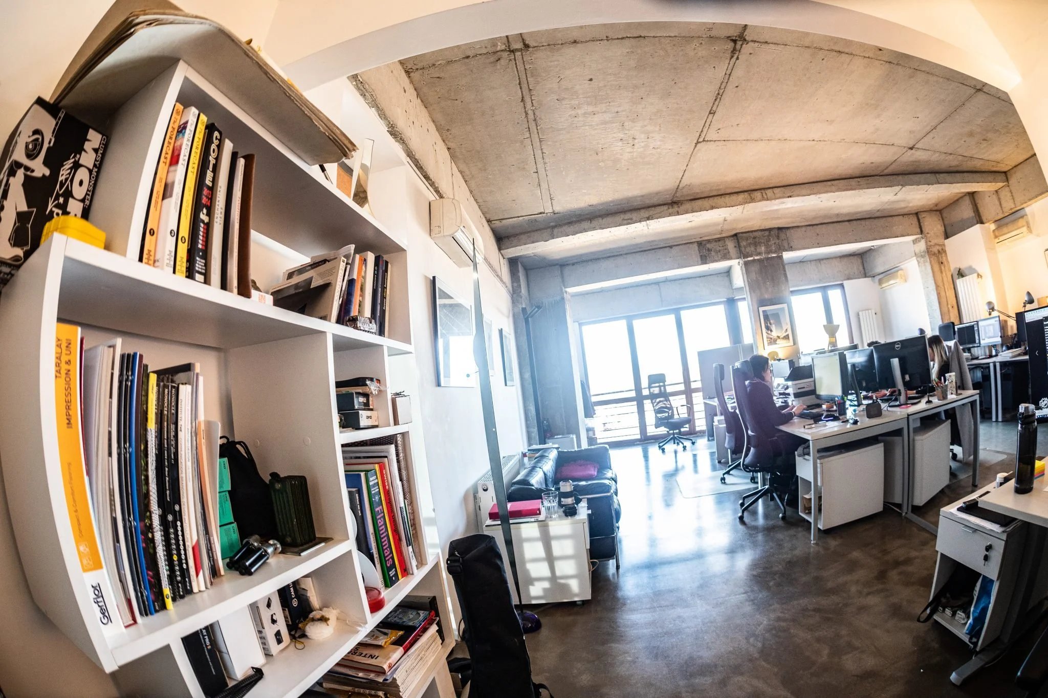 Modern open-plan office with ergonomic chairs, desks, computers, and large windows letting in natural light. Decor includes framed artwork, a black leather couch, and a white bookshelf with books and office supplies. Concrete ceiling and polished floor contribute to industrial style.