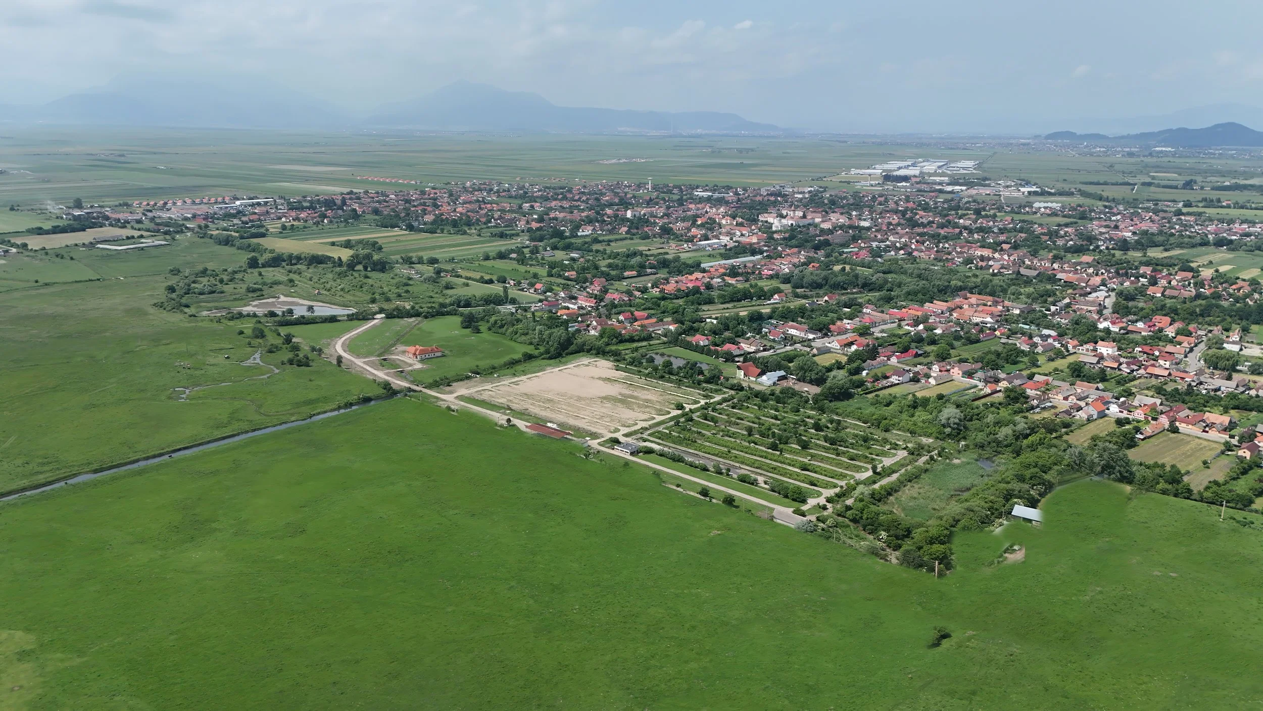 An aerial view of a rural town with green fields, small roads, and houses with red roofs, extending into a distant cityscape with mountains in the background.