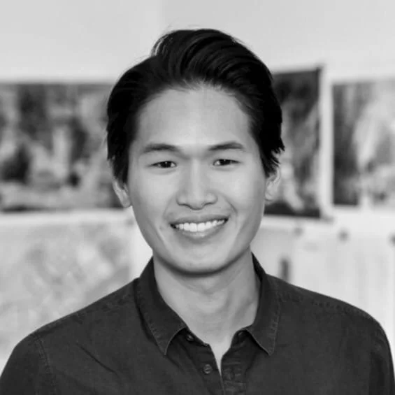 A young man with short dark hair, smiling, wearing a dark button-up shirt, in front of blurred art or photos.