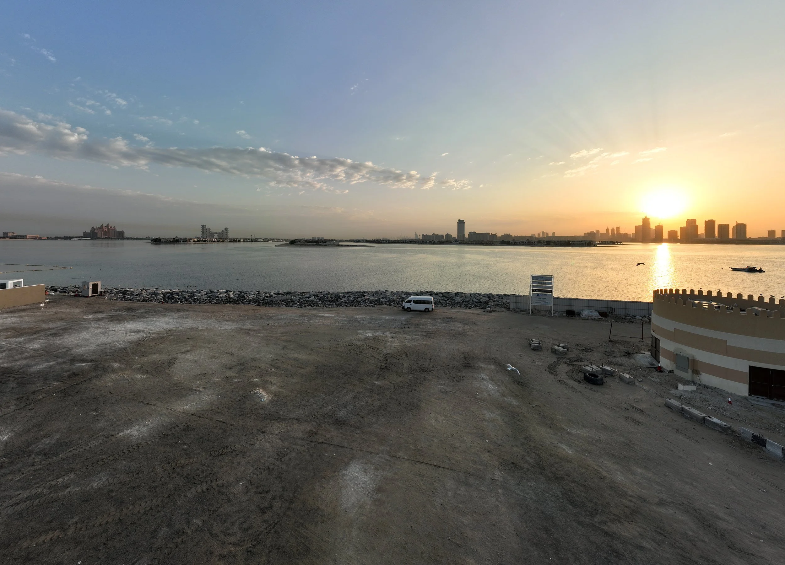 A sunset over a city skyline across a water body, with a construction site in the foreground and a white van parked near the water.