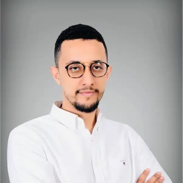 Portrait of a man with short dark hair, glasses, a beard, wearing a white collared shirt, standing against a plain gray background.