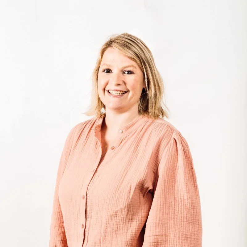 A smiling woman with shoulder-length blonde hair wearing a peach-colored blouse, standing against a plain white background.