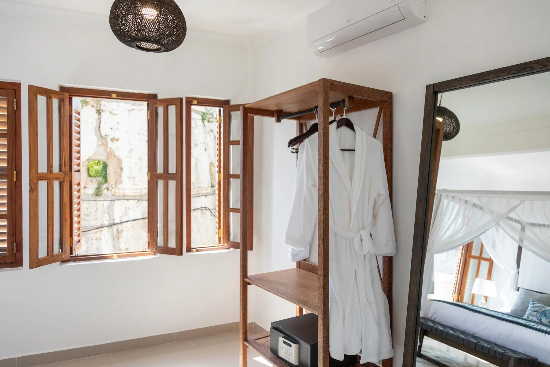 A room with open wooden window shutters, a standing wooden clothing rack with a white bathrobe, a small safe, a mirror with a dark frame, and a wall-mounted air conditioning unit.