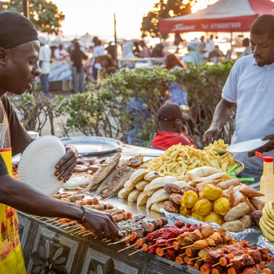 People at a street food stall with skewered meats, fried snacks, French fries, and other dishes, with outdoor setting and other food vendors in the background during sunset, Forodhani Market, Stone Town, Zanzibar.