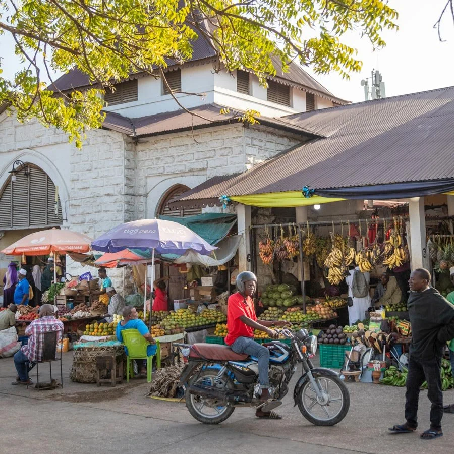 Darajani Market selling fruits with vendors, shoppers, a man on a motorcycle, and a building in the background, Stone Town, Zanzibar.