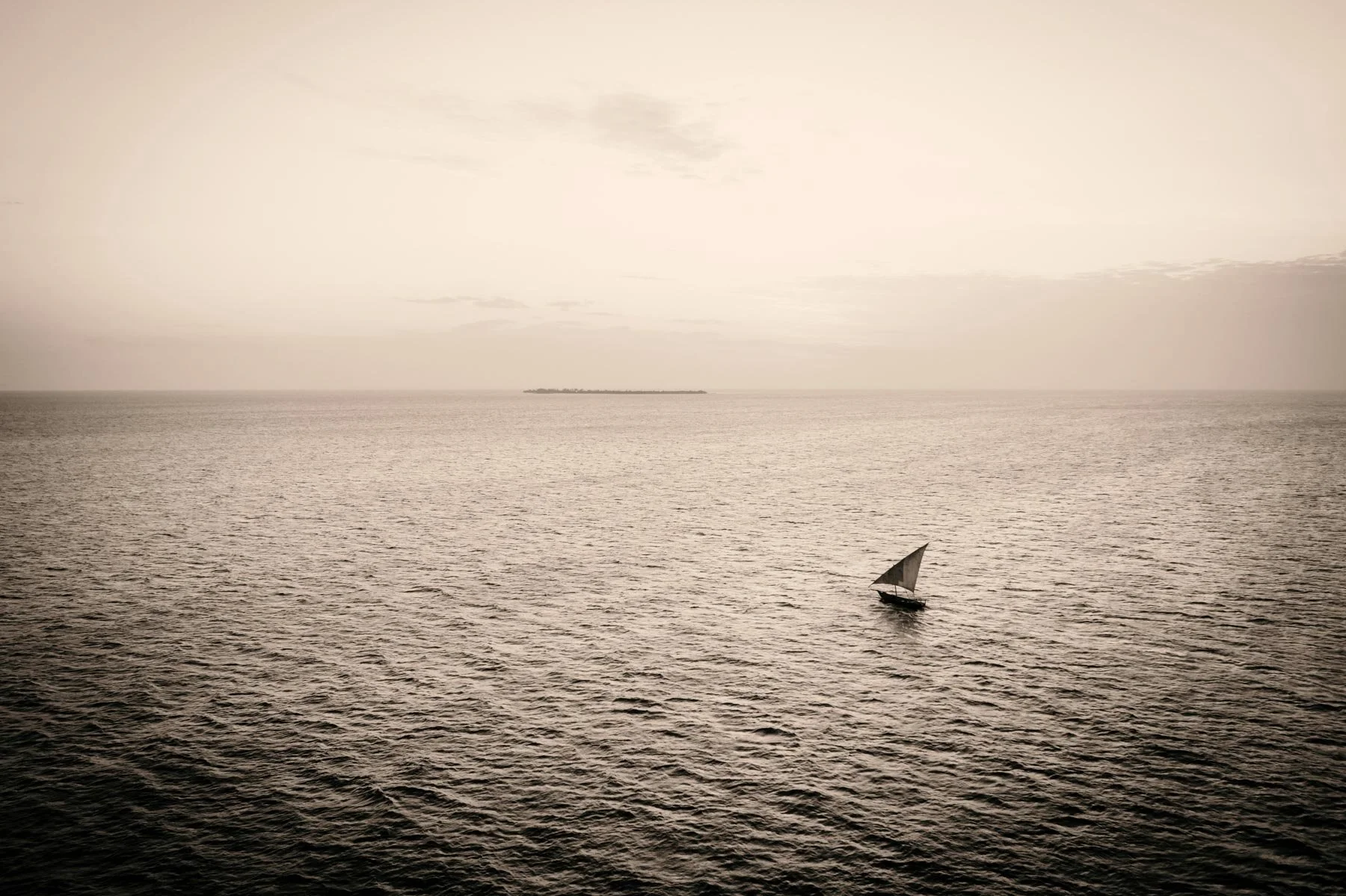 A small sailboat with a dark hull and a white sail navigating calm waters near sunset, with a distant landmass on the horizon.