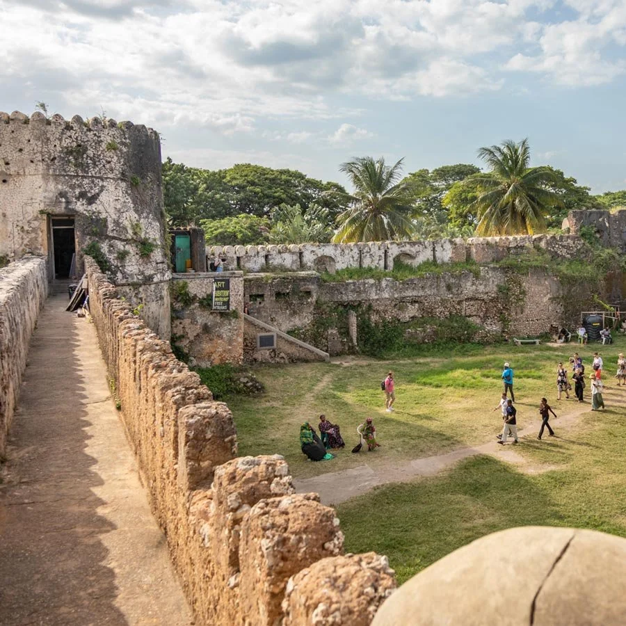 View of the Old Fort with a walking path on the left, a grassy courtyard with people, and palm trees in the background, Stone Town, Zanzibar.
