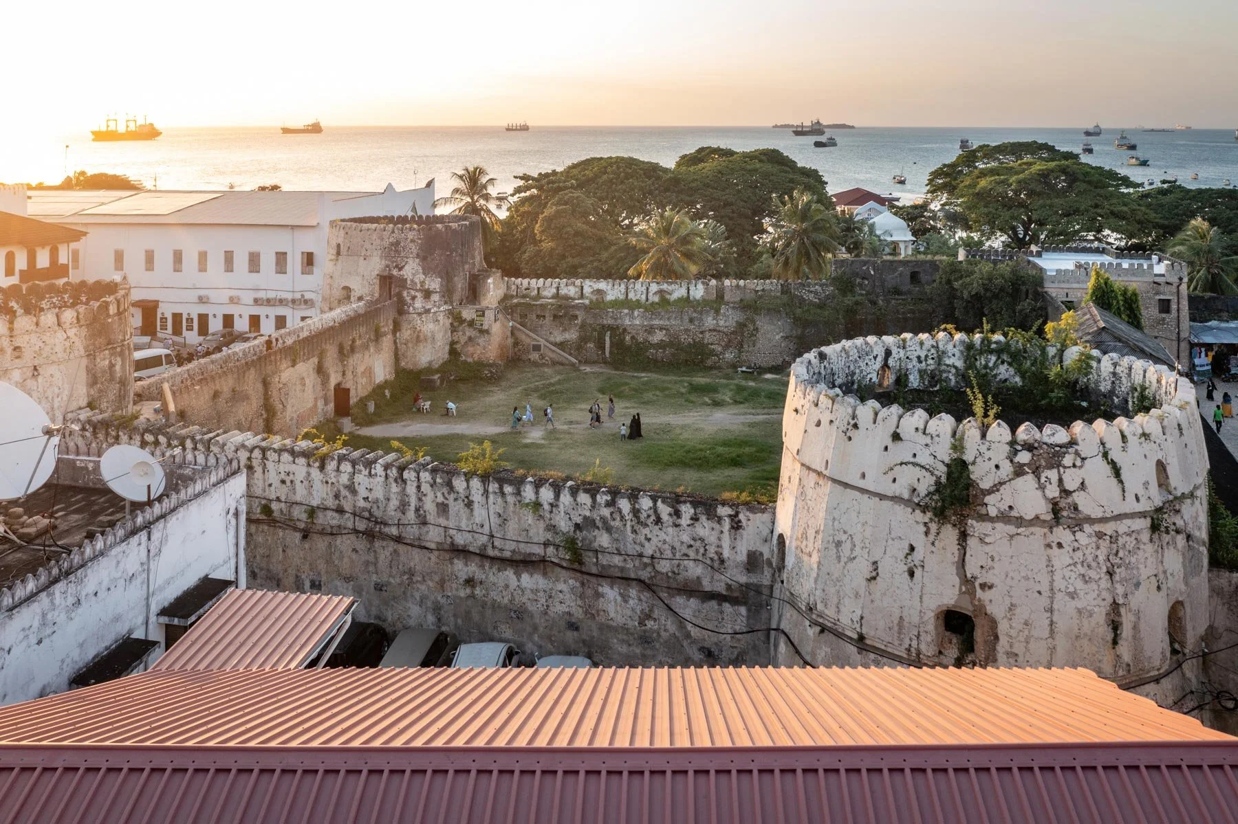 Aerial view of a historic lighthouse and surrounding fortress walls overlooking a colonial city with a harbor, ships, and palm trees at sunset.