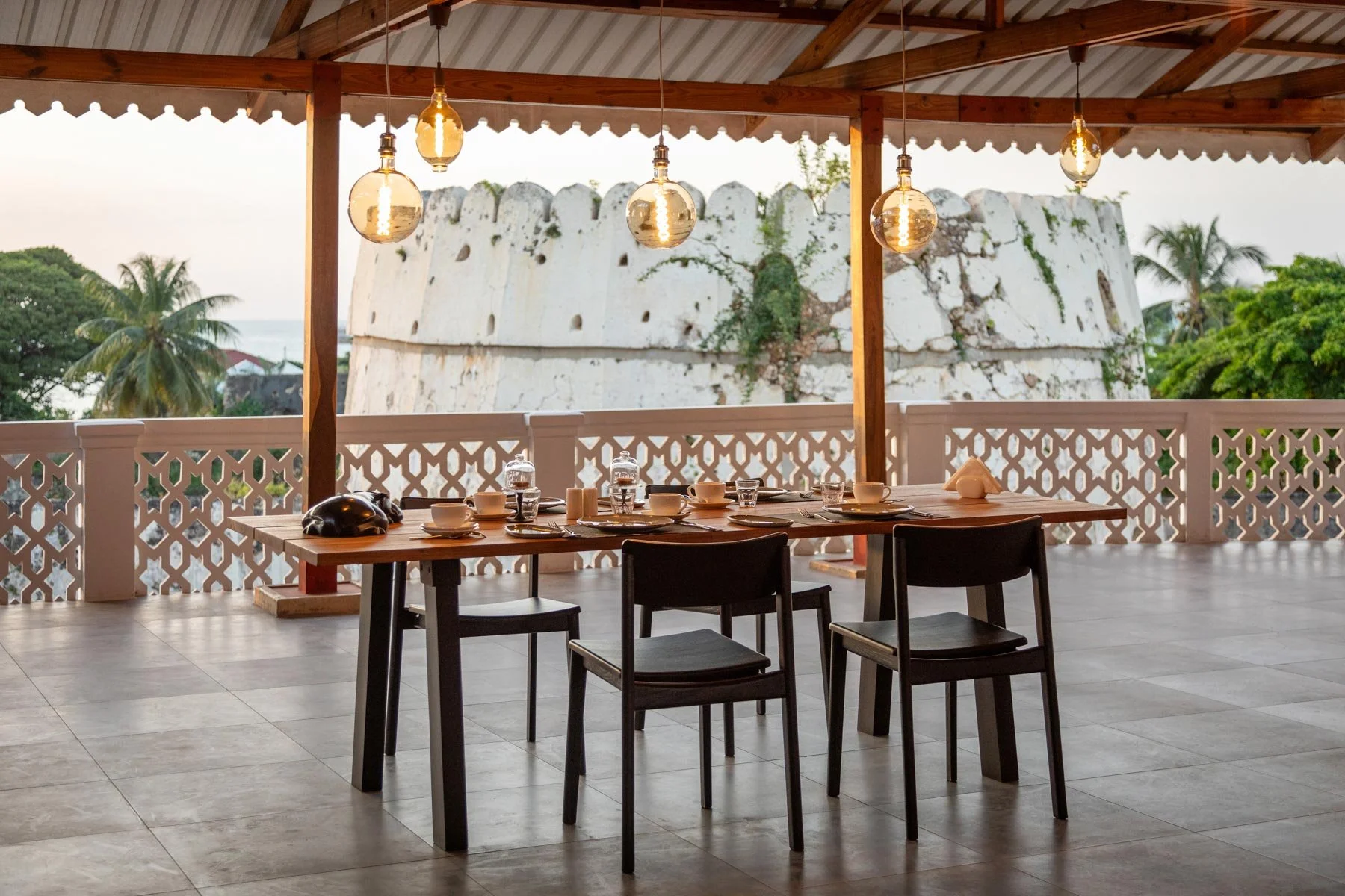 An outdoor dining area with a wooden table set for a meal, surrounded by black chairs, under a wooden roof with hanging pendant lights. In the background, a white stone structure and tropical greenery are visible.