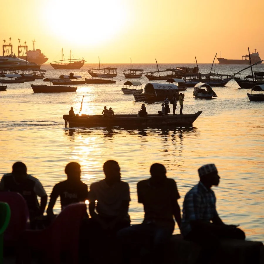 Forodhani Gardens at Sunset - silhouettes of five people sitting and standing by the water at sunset, with numerous boats anchored in the bay and a large ship in the distance. Stone Town, Zanzibar.
