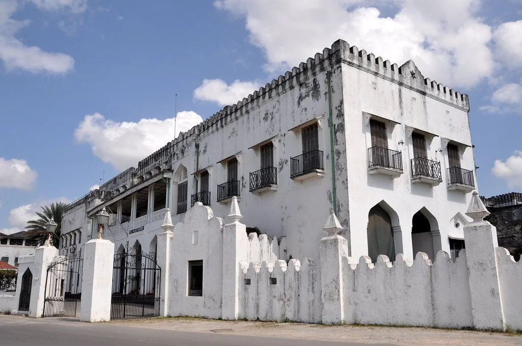 The Sultan's Palace - a white historic building with arched doorways, small balconies, and a decorative battlement-style roofline, surrounded by a white wall, under a partly cloudy sky. Stone Town, Zanzibar.