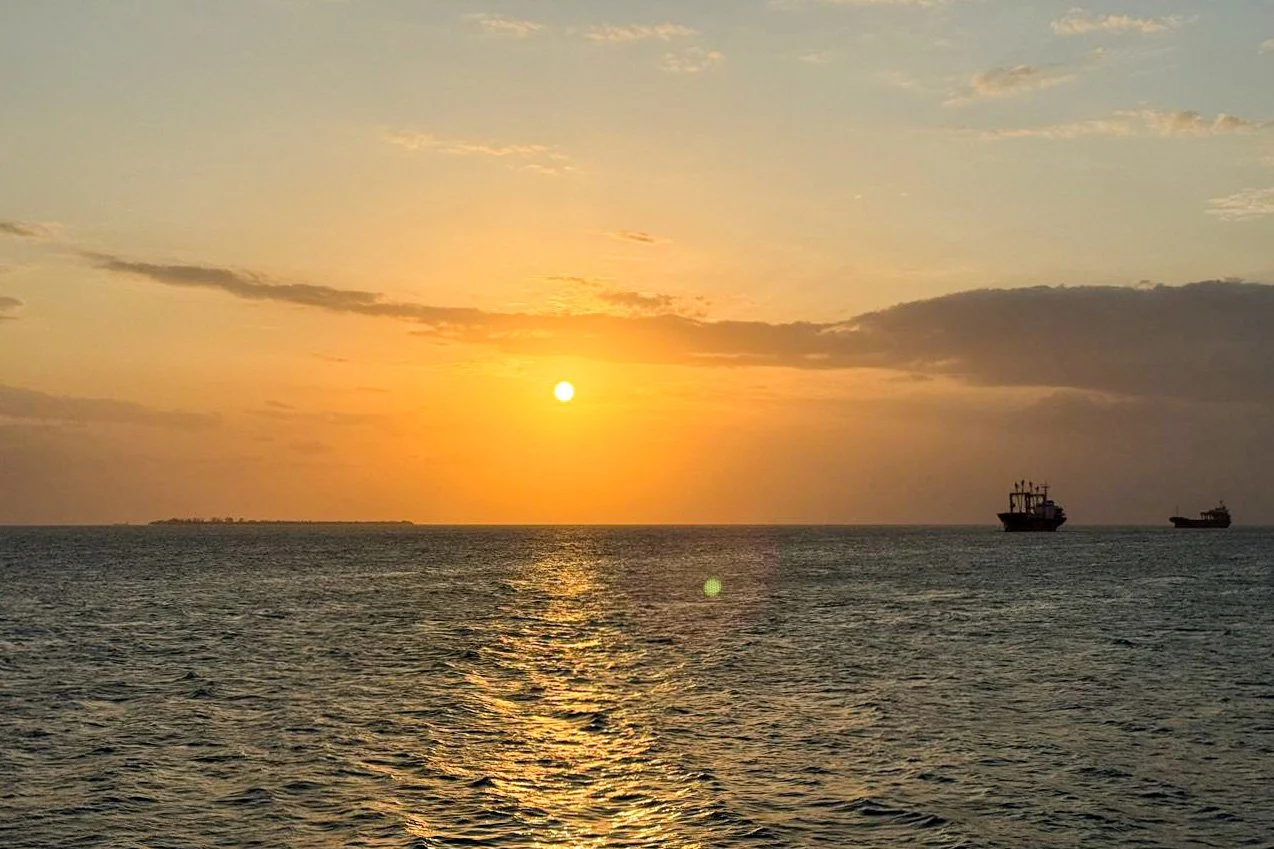 Sunset over the ocean with two ships silhouetted against the sky.