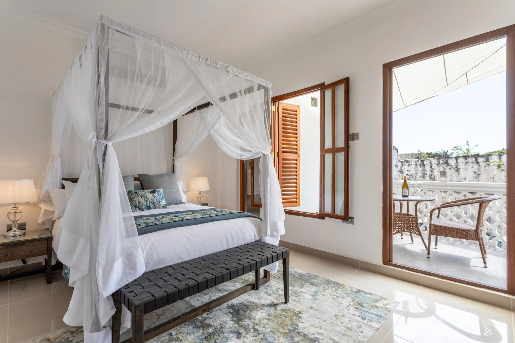 A bedroom with a four-poster bed covered with white canopy netting, two bedside lamps, and a bench at the foot of the bed. There is an open door leading to a balcony with outdoor chairs, a table with a wine bottle, and a scenic view.