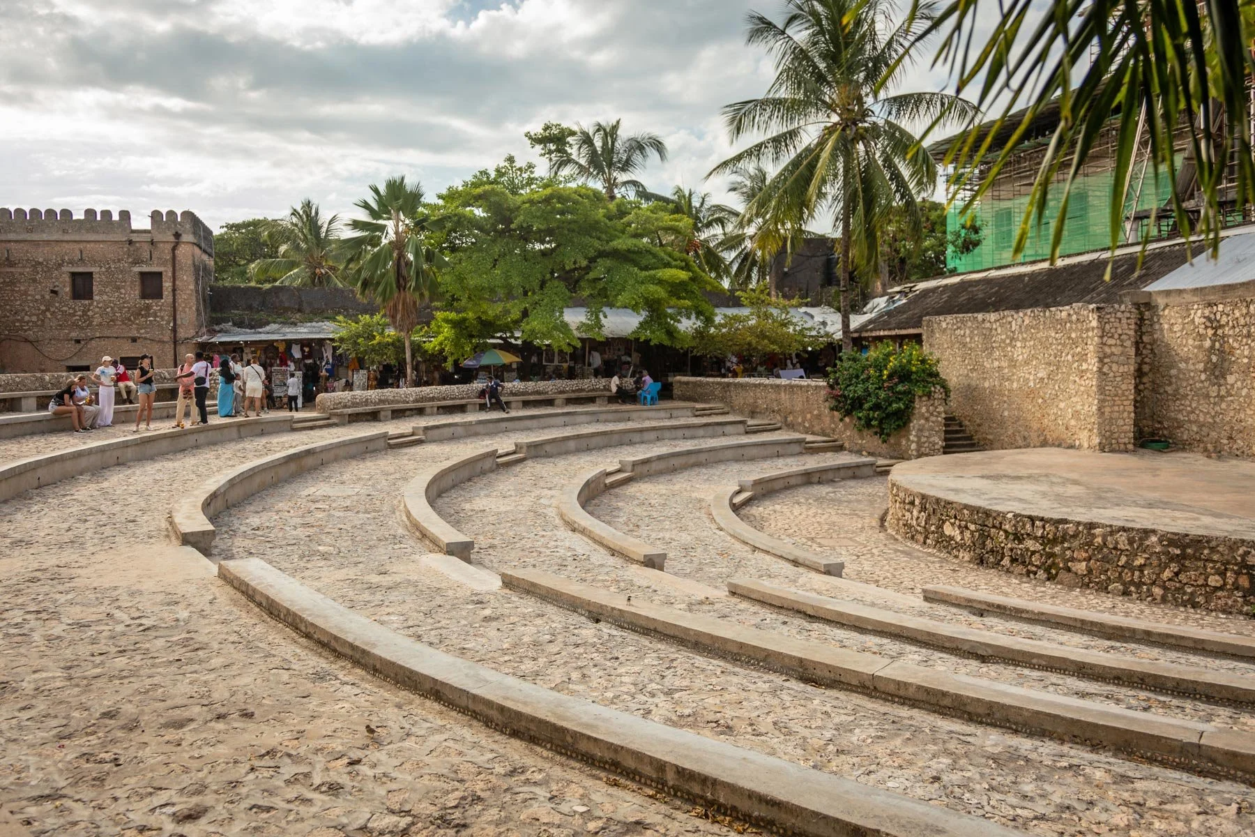 Open-air amphitheater of the Old Fort with curved stone seating surrounded by trees and historic stone buildings in a lively outdoor market. Stone Town, Zanzibar.