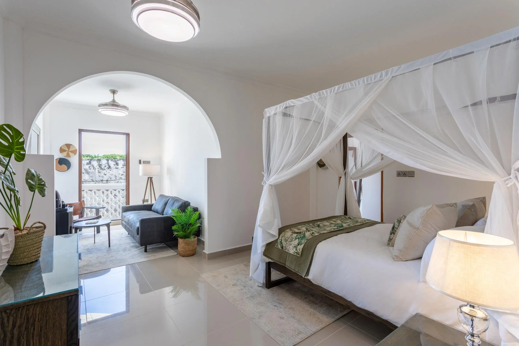 A bedroom with a four-poster bed draped in white canopy curtains, beige bedding, and tropical-themed accents. A living area with a black sofa, potted plants, and decorative wall art is visible through an arched opening.