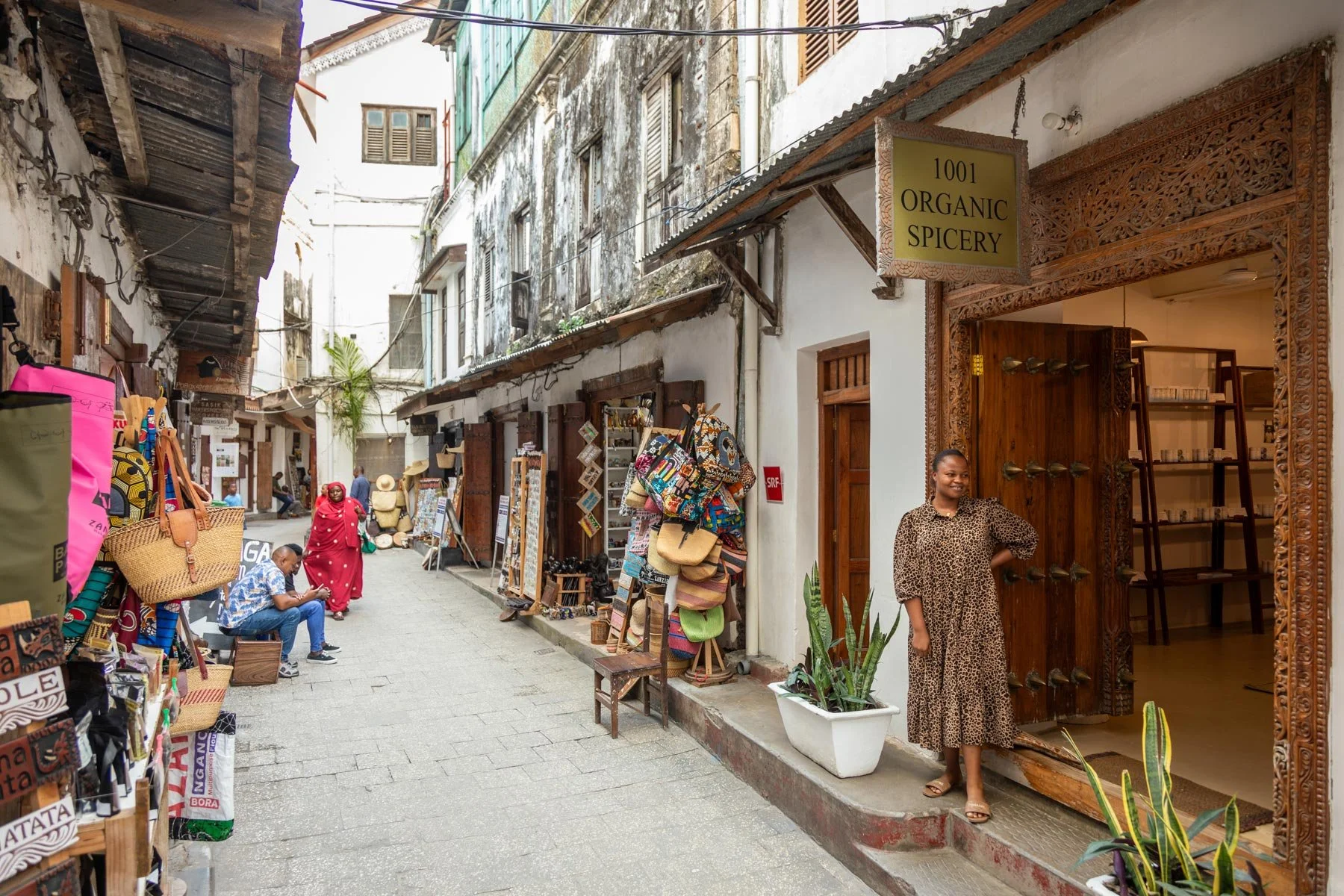 Street scene with small shops and vendors, a woman standing at the entrance of a shop with a sign that reads '1001 Organic Spicery', and people walking or sitting on the sidewalk.