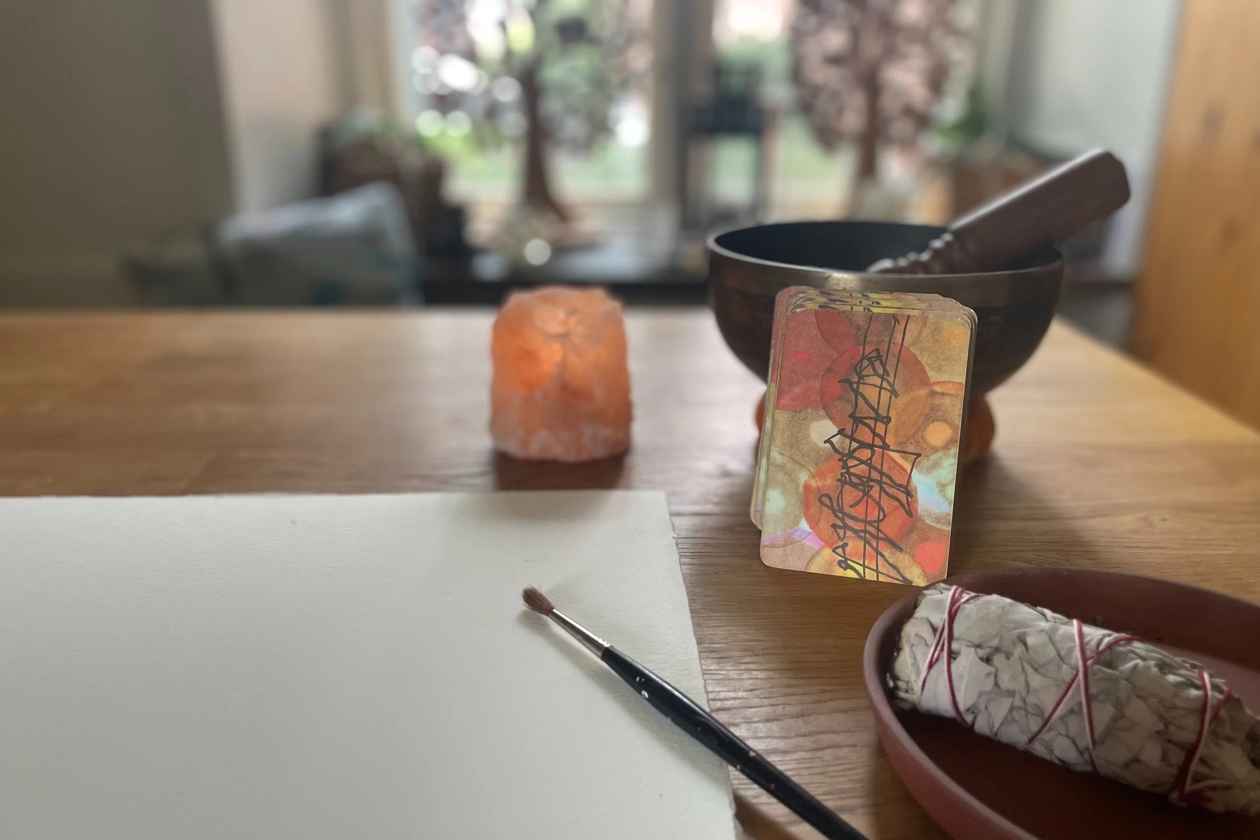 Konstnärs-material på ett bord inklusive en tom vit canvas, en pensel, en saltstenslykta, en tarotlek, en klangskål, samt ett knippe salvia