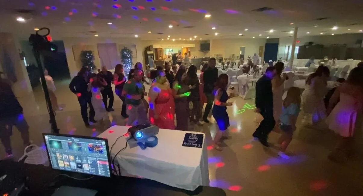 People dancing in a large banquet hall with white tablecloth-covered tables, string lights, and balloons, illuminated by colorful disco lights.