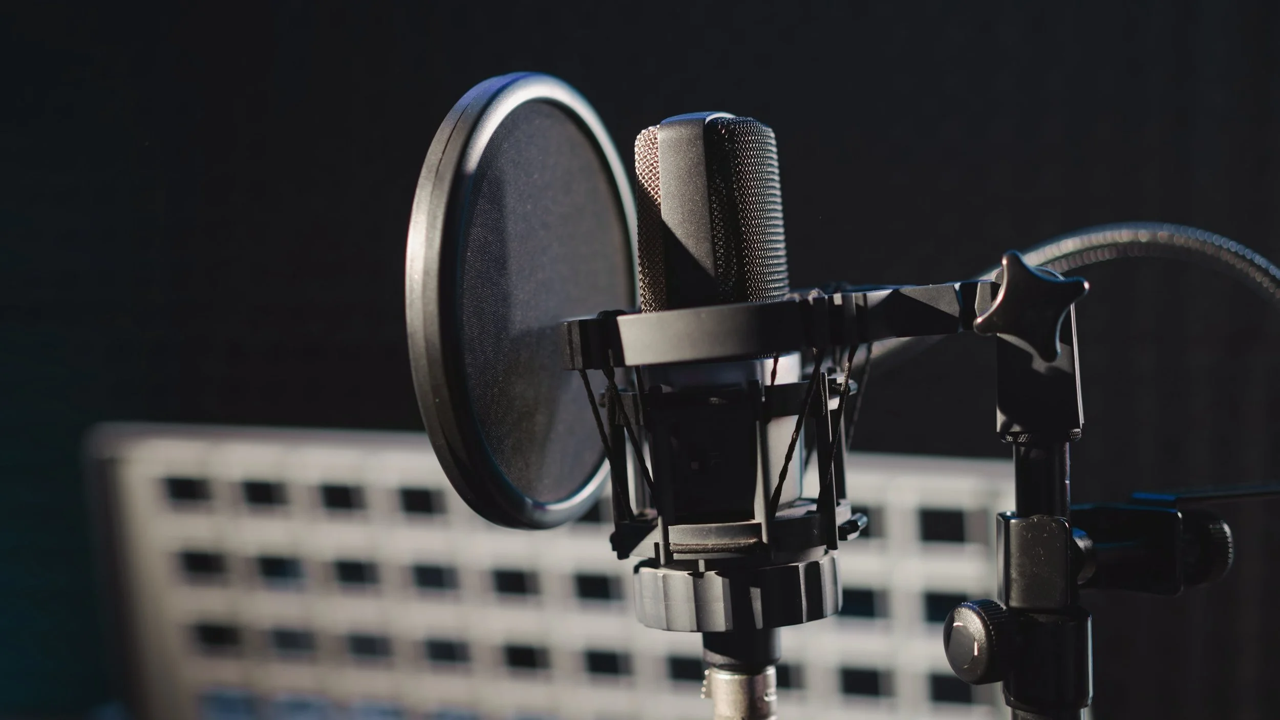 Close-up of a professional microphone with a pop filter, set up in a recording studio, with a black soundproof background and grid-like panel in the background.
