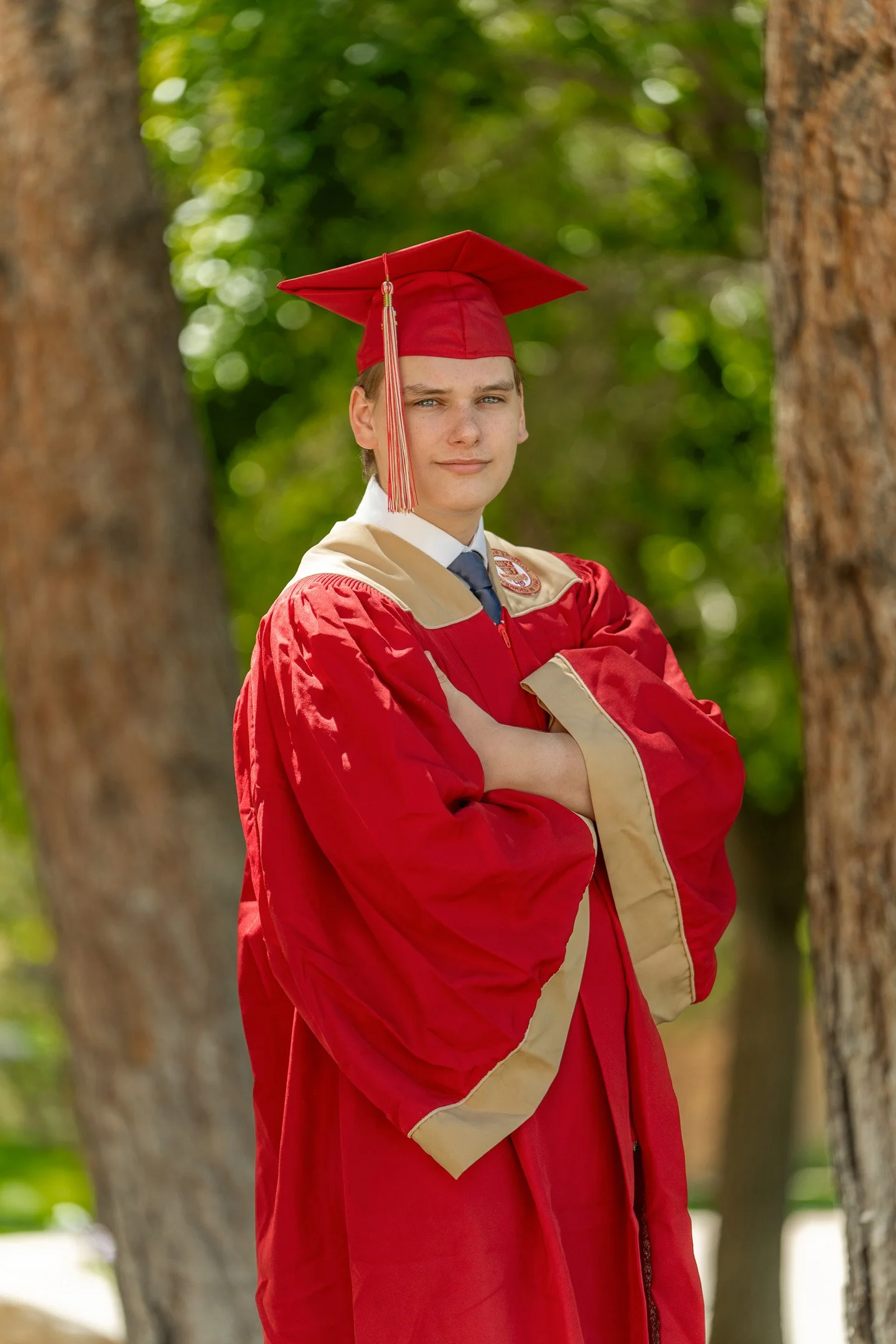A high school senior in cap and gown standing, posing for their senior portrait in a grass field amongst the trees as a bokeh background