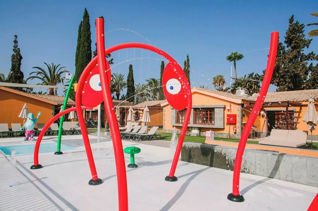 Juegos de agua en una piscina con estructuras rojas con caras, en un área de recreo con edificios de color tierra y palmeras al fondo.