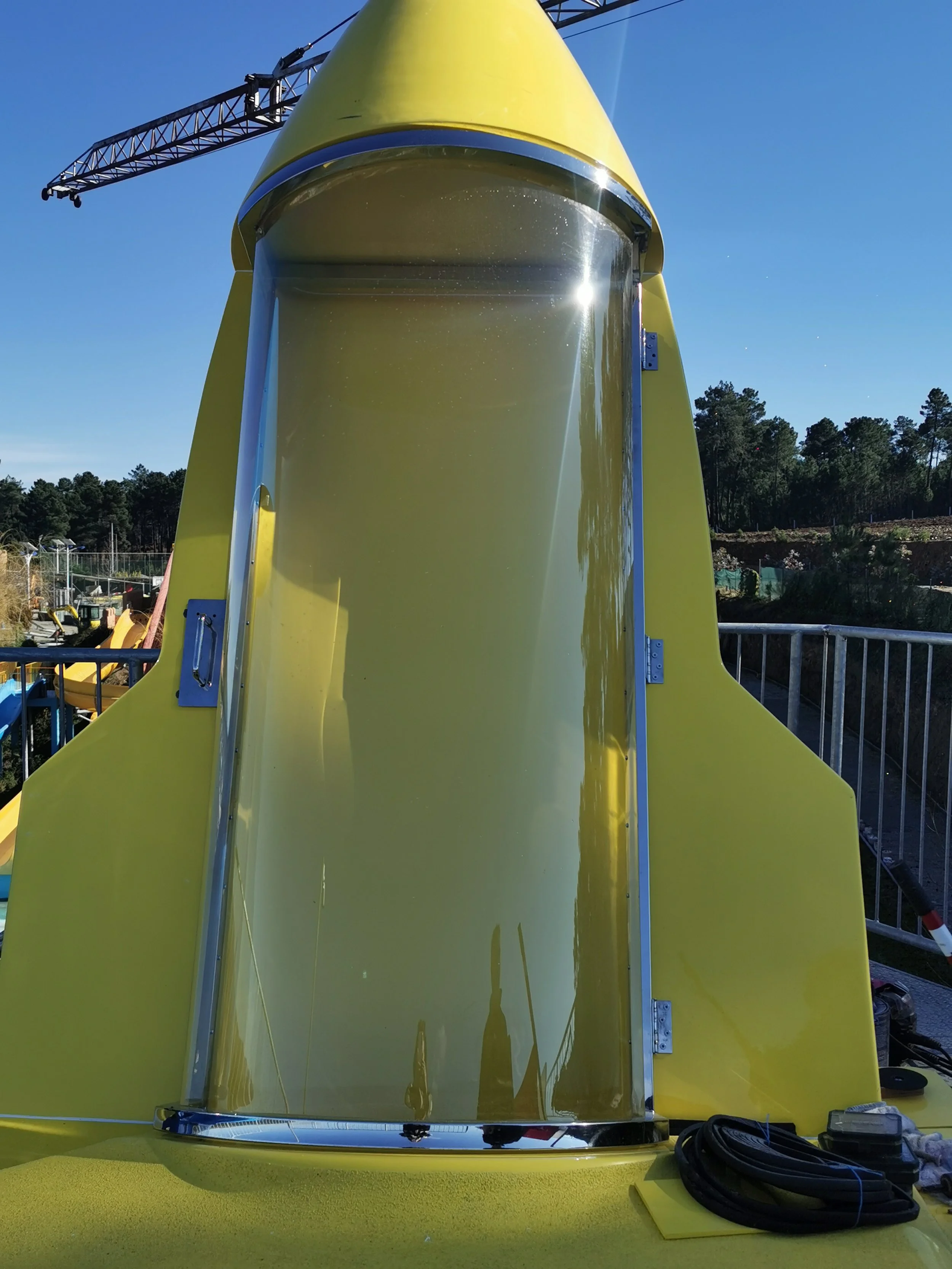 Vista frontal de una atracción de parque acuático tipo tobogán amarillo con tobogán de vidrio transparente, en exteriores con cielo azul y árboles de fondo.