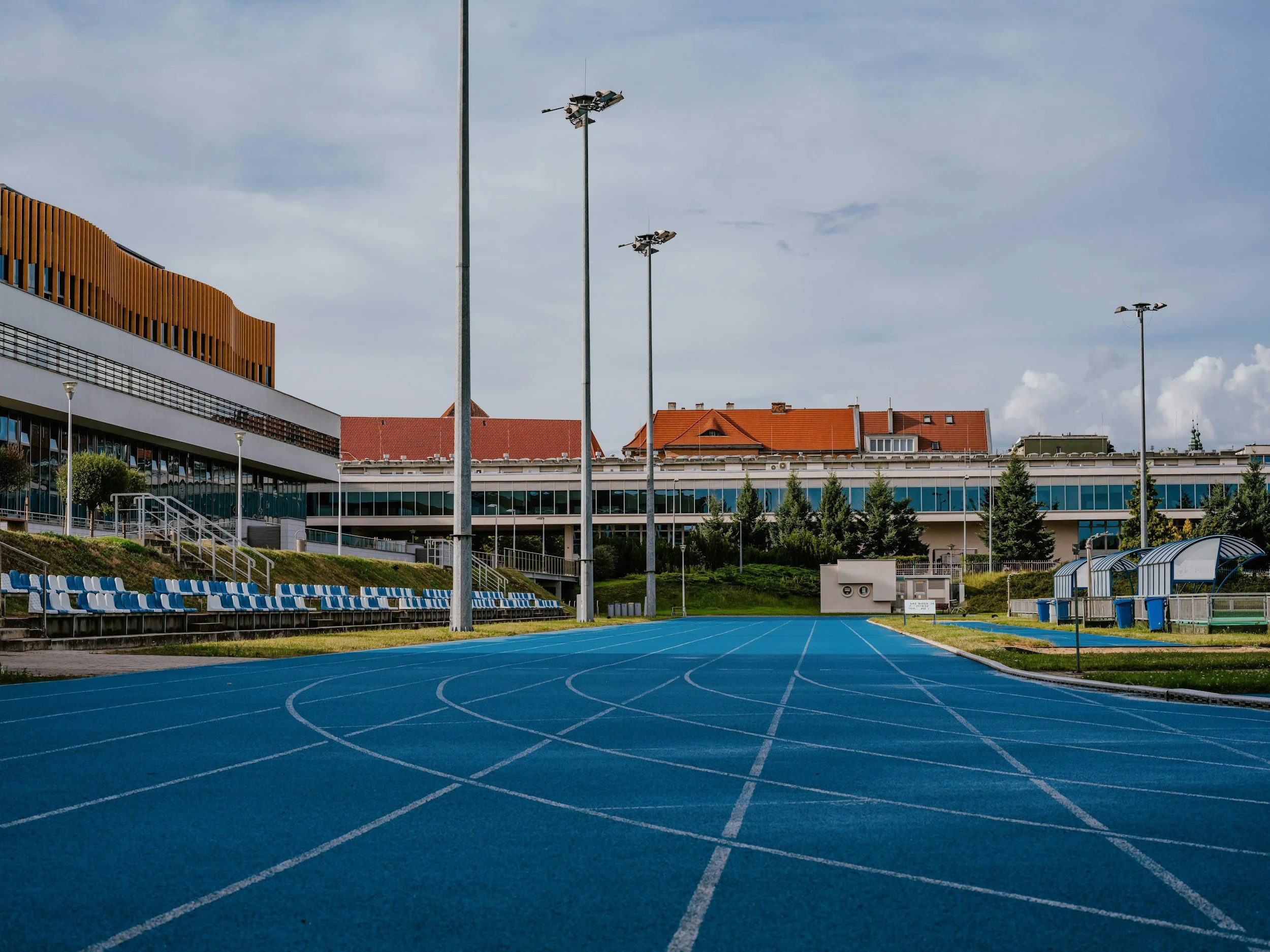 Carril de atletismo azul en estadio al aire libre, con gradas y edificios en el fondo, y luminarias altas.