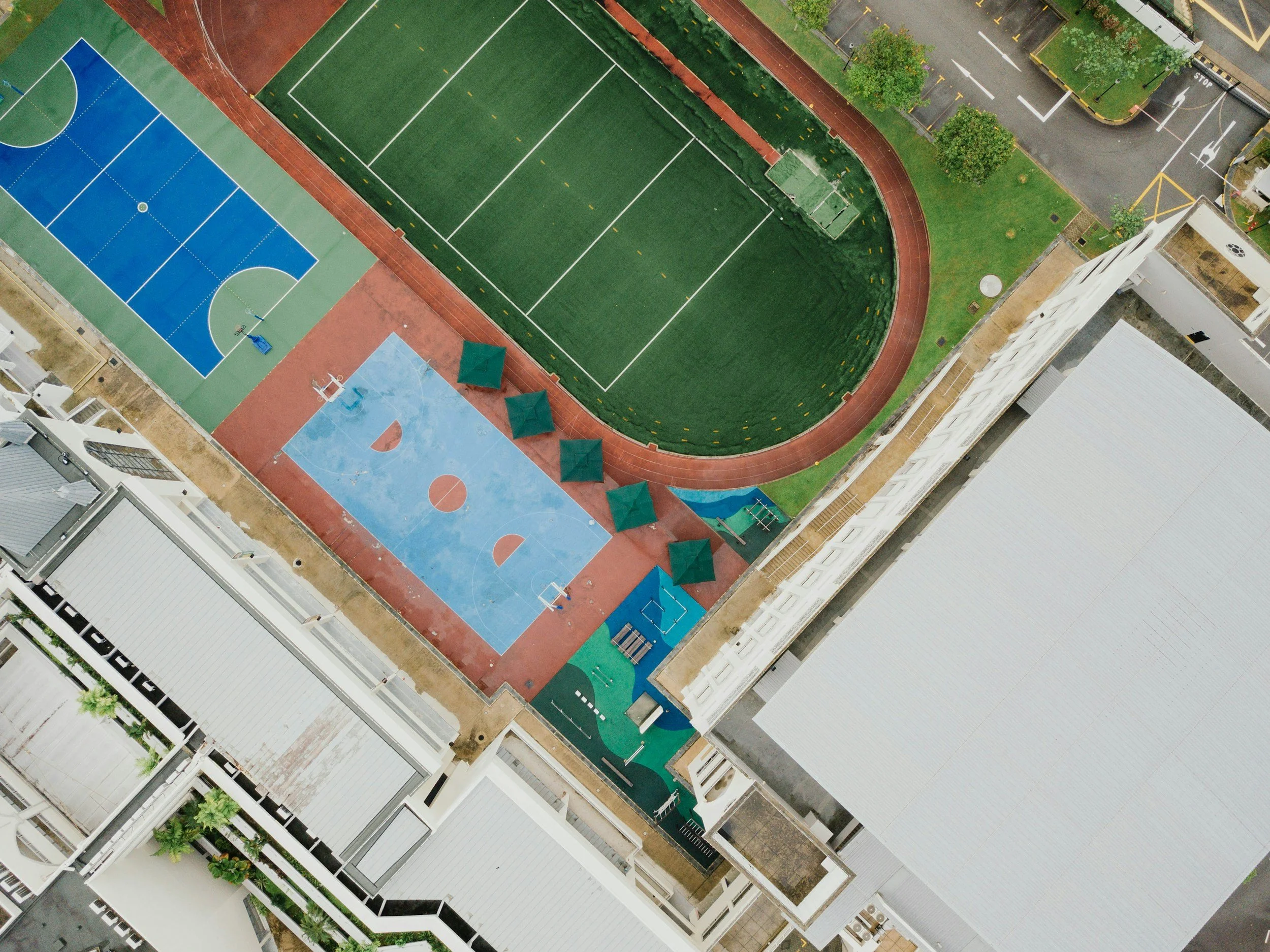 Vista aérea de una área recreativa en una ciudad, mostrando un campo de fútbol, una pista de atletismo, varias canchas de baloncesto, áreas de sombra con sombrillas verdes y un parque infantil en un entorno urbano con edificios y calles