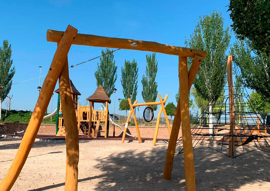 Parque infantil con columpios de madera y estructuras de escalada, rodeado de árboles bajo un cielo despejado.