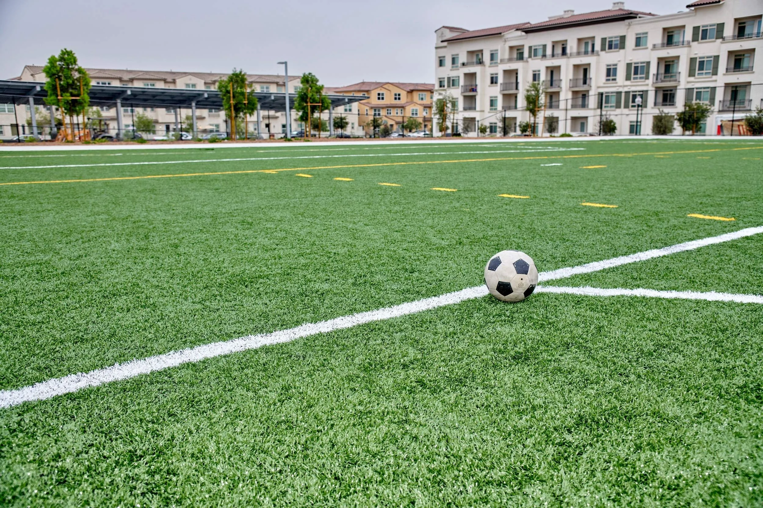 Balón de fútbol en un campo de césped artificial con edificios residenciales en el fondo.