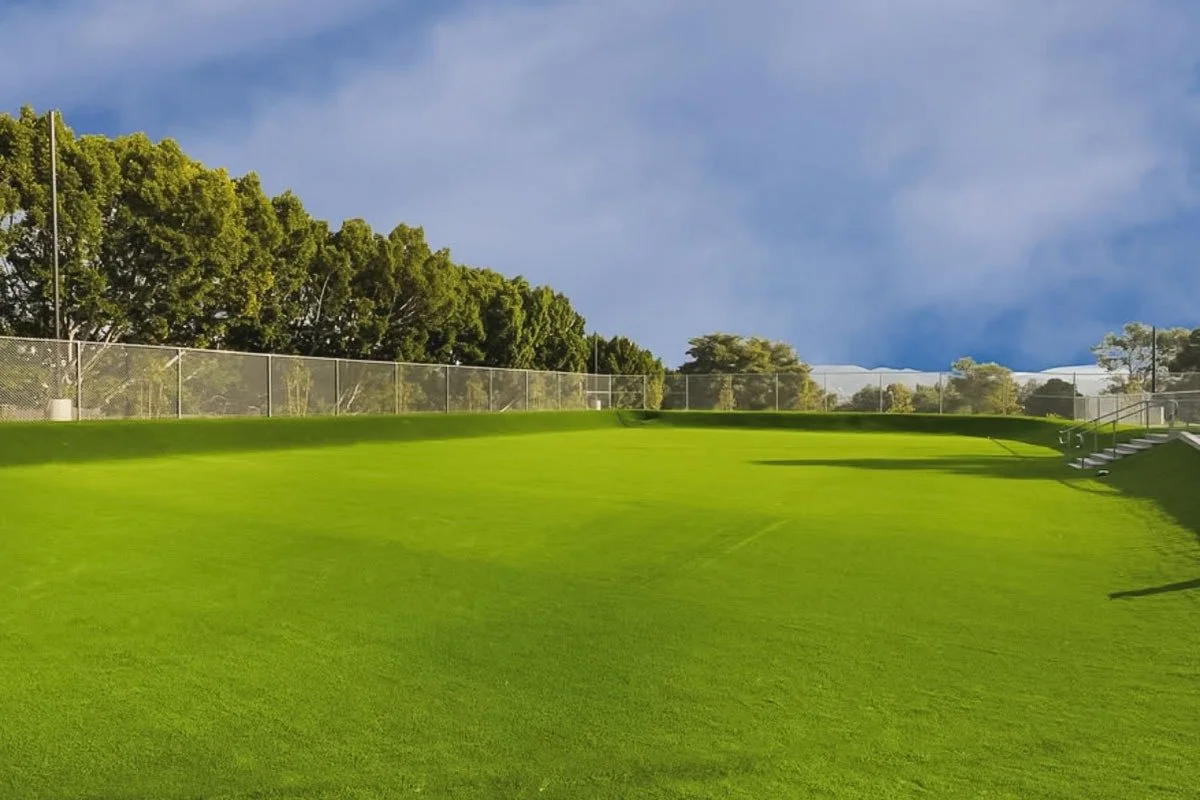 Campo de golf con césped verde, vallas de protección y árboles al fondo, cielo parcialmente nublado.