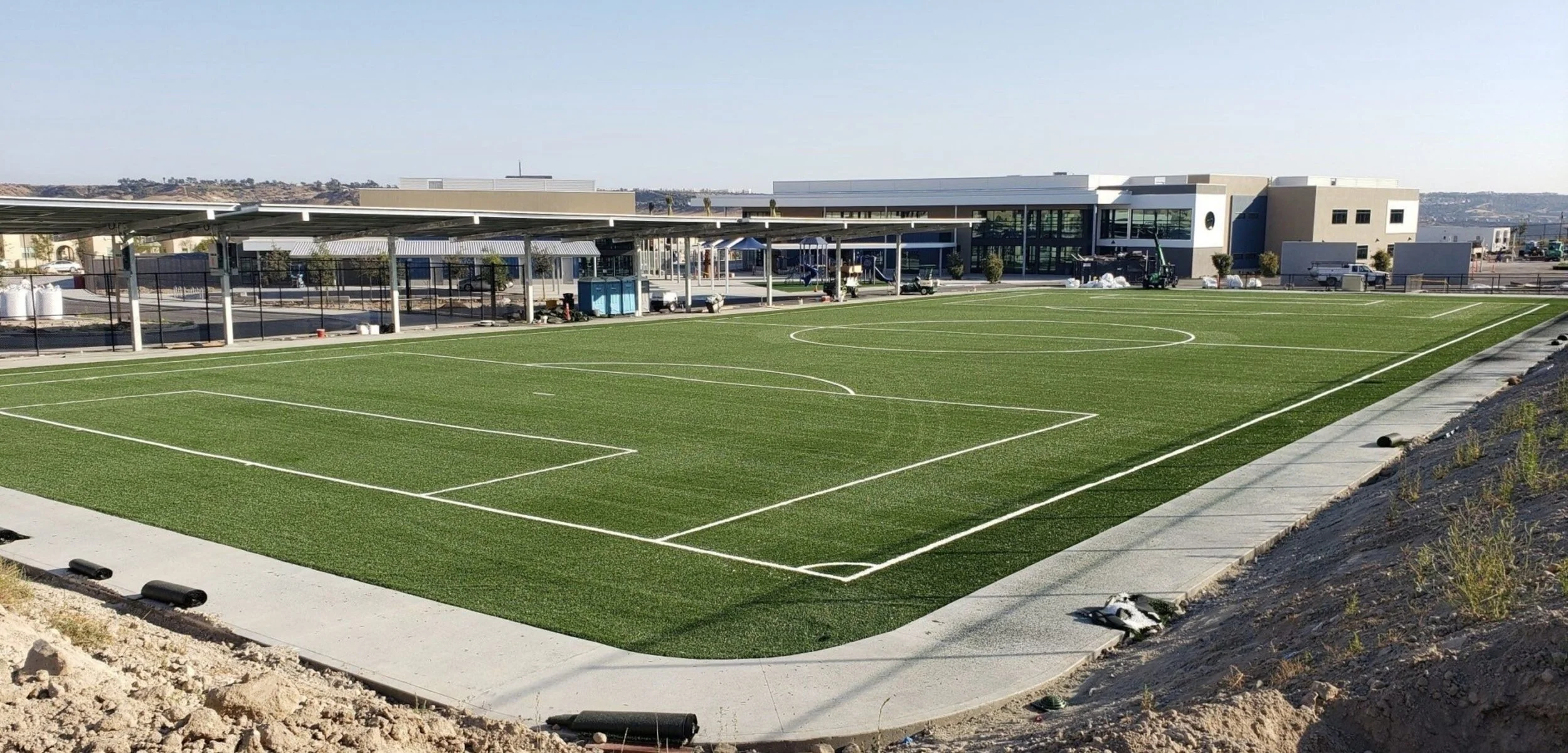 Campo de fútbol con grama artificial y líneas blancas en las límites y marcas del campo, edificio moderno en el fondo y terreno arenoso en el primer plano.