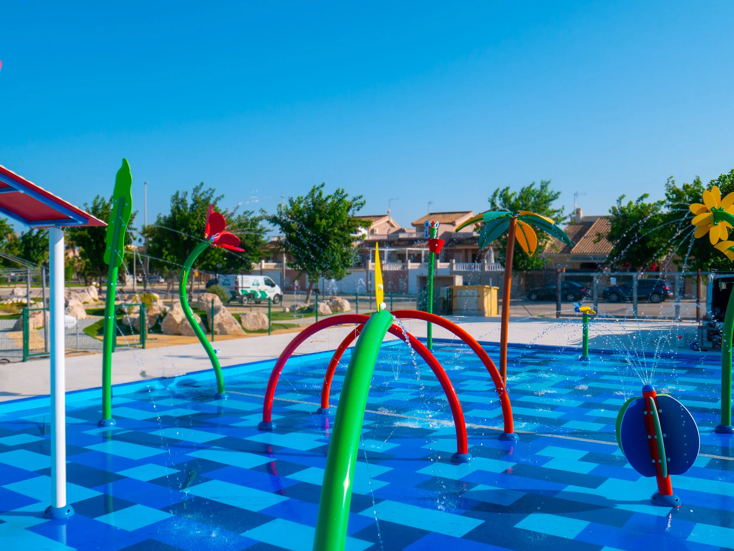 Parque infantil con fuentes de agua y estructuras de juego en exteriores, con casas residenciales y árboles en el fondo.