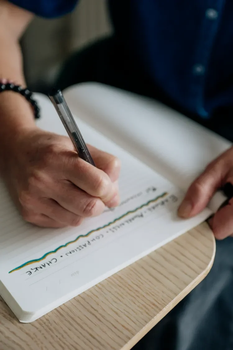 A therapist writing notes in a notebook with a black pen, sitting at a wooden table.