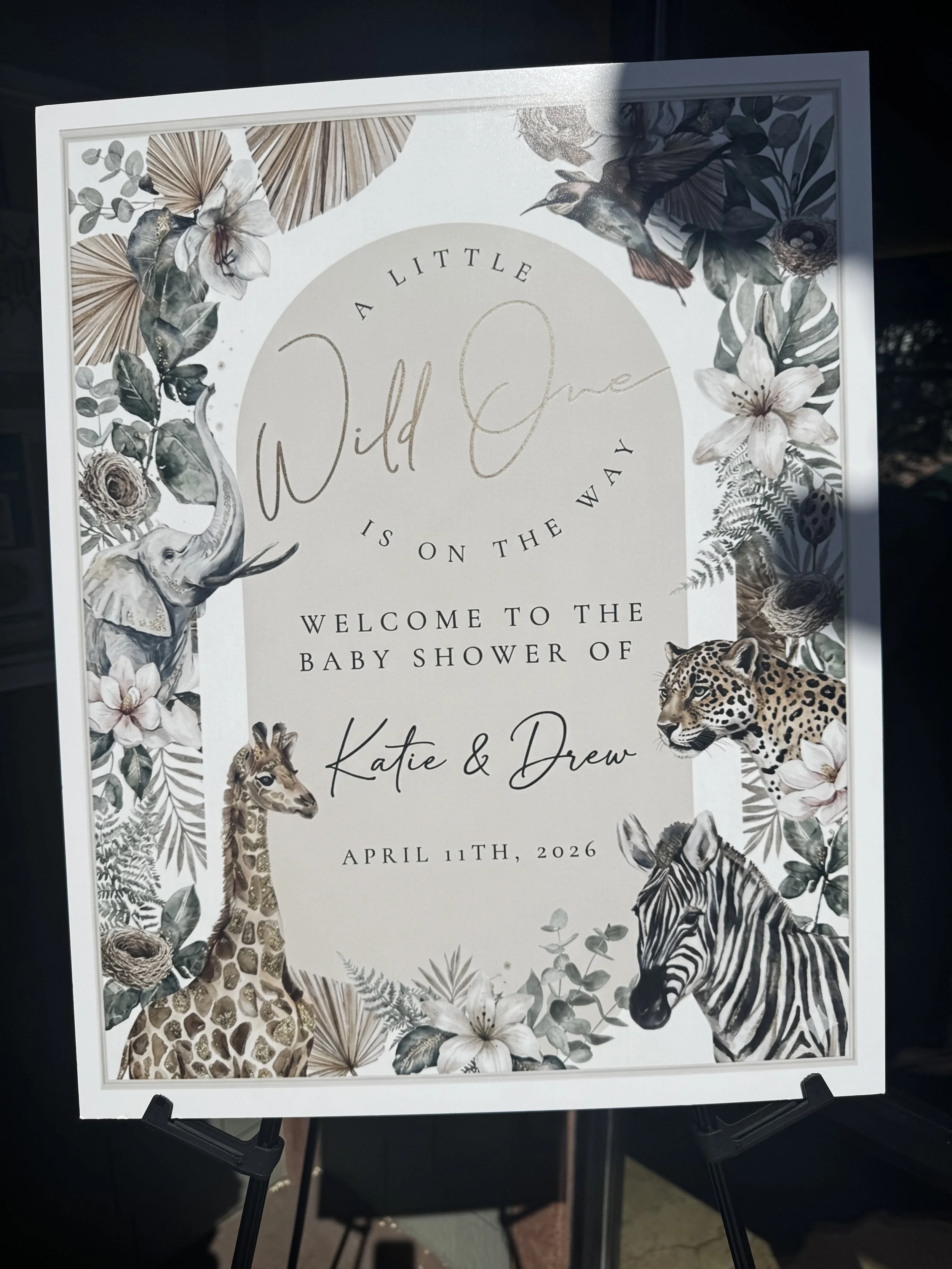 Baby shower invitation with animal illustrations, including a giraffe, zebra, elephant, cheetah, and various birds, surrounded by leaves and flowers, announcing the baby shower of Katie and Drew on April 11th, 2026.