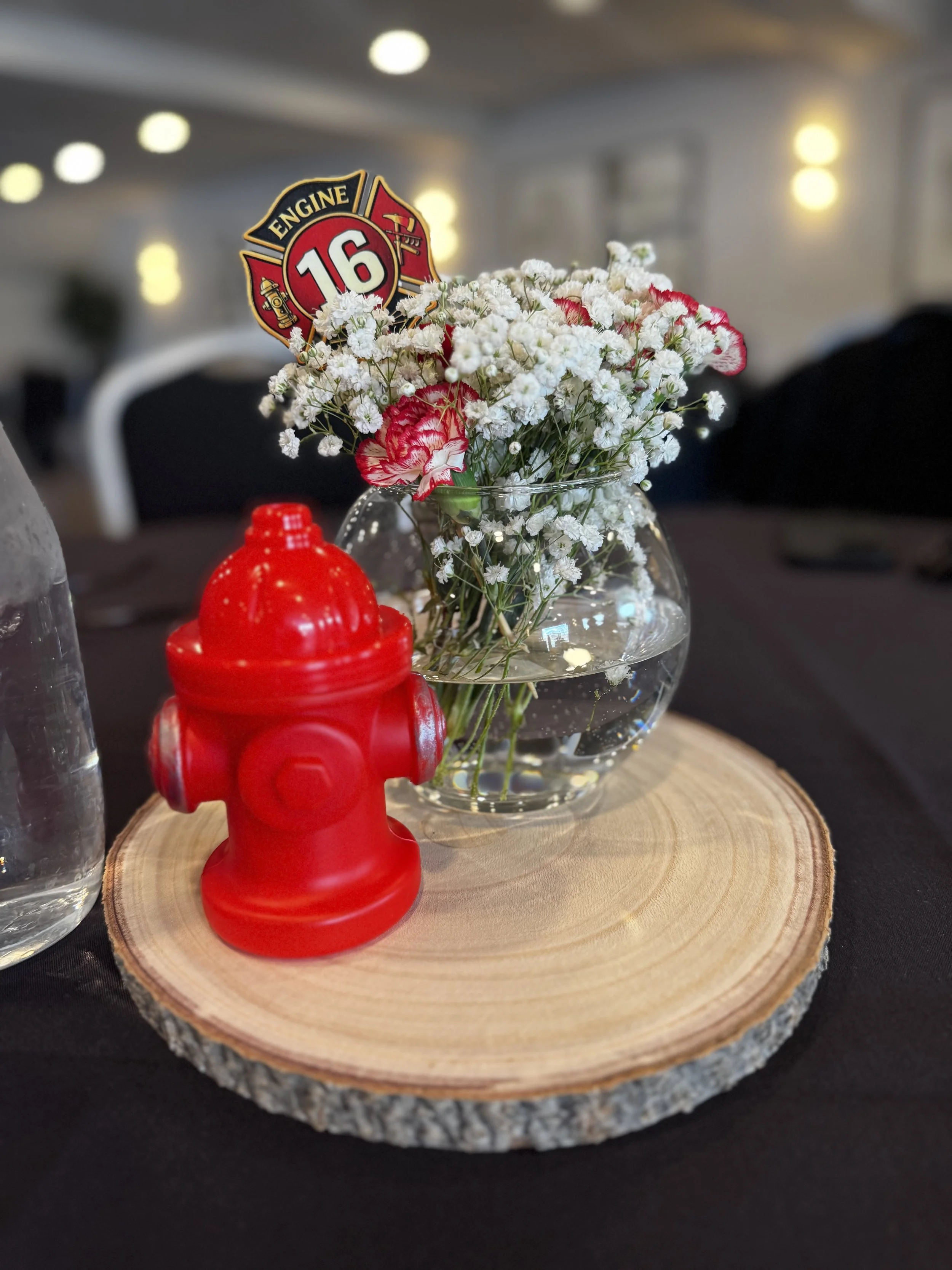 A glass bowl with white and pink flowers, a fire hydrant-shaped paper decoration, and a red object on a round wood slice.