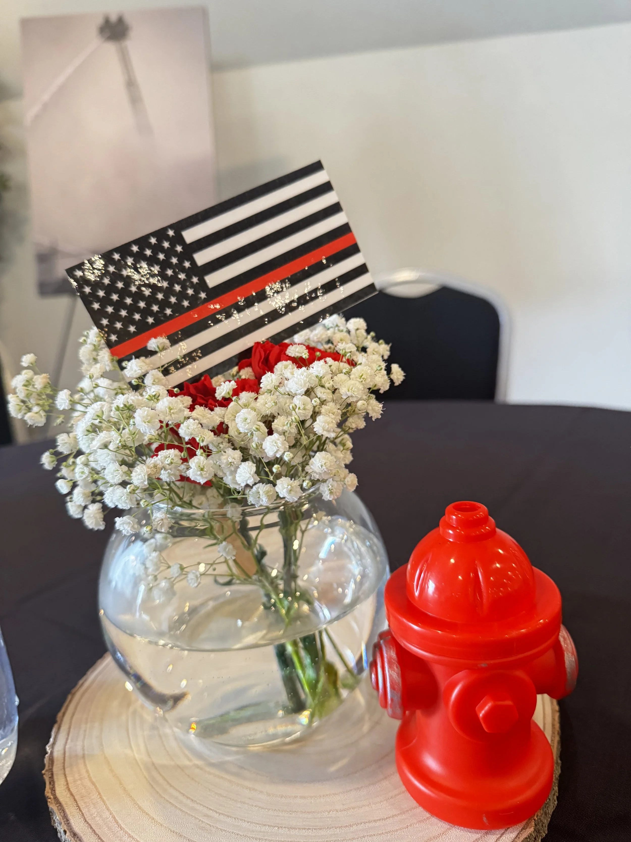 Glass vase with white baby's breath and red roses, American flag themed card leaning against the flowers, red sippy cup on a wooden round table, with a blurred chair and wall in the background.