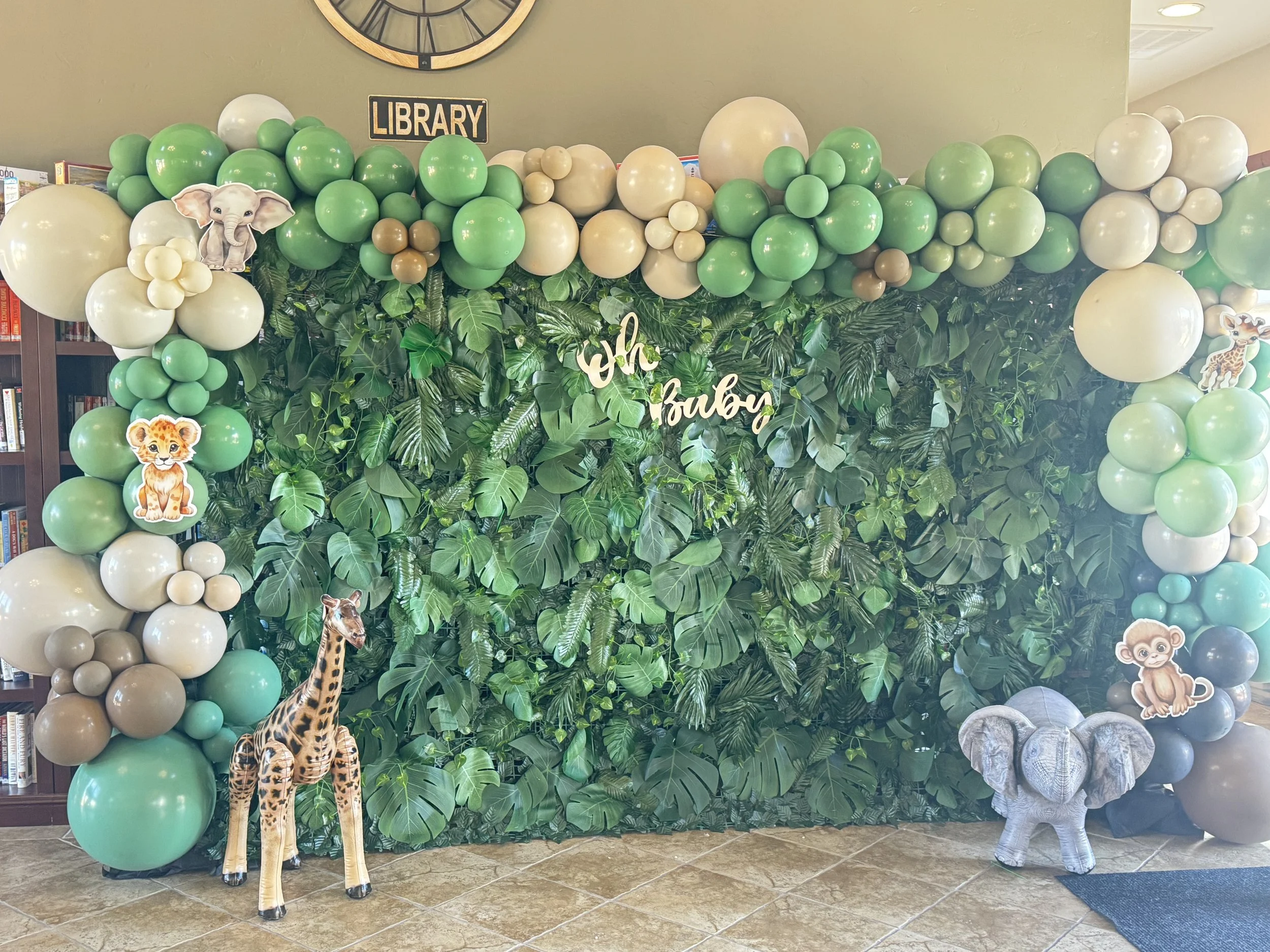 Decorative jungle-themed display with green, beige, and white balloons, artificial green leaves, and cardboard cutouts of a giraffe, elephant, monkey, and zebra.