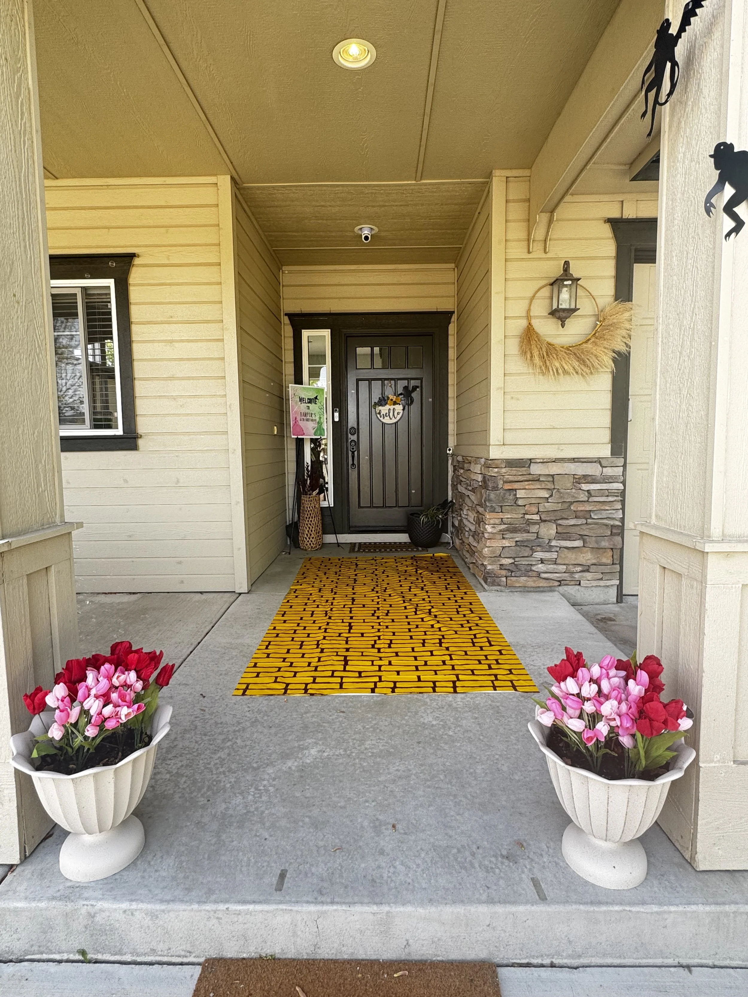 Entryway of a house with a black front door, a yellow brick-patterned doormat, two white planters with pink and red flowers, decorative black animal silhouettes on the wall, and a hanging floral wreath.