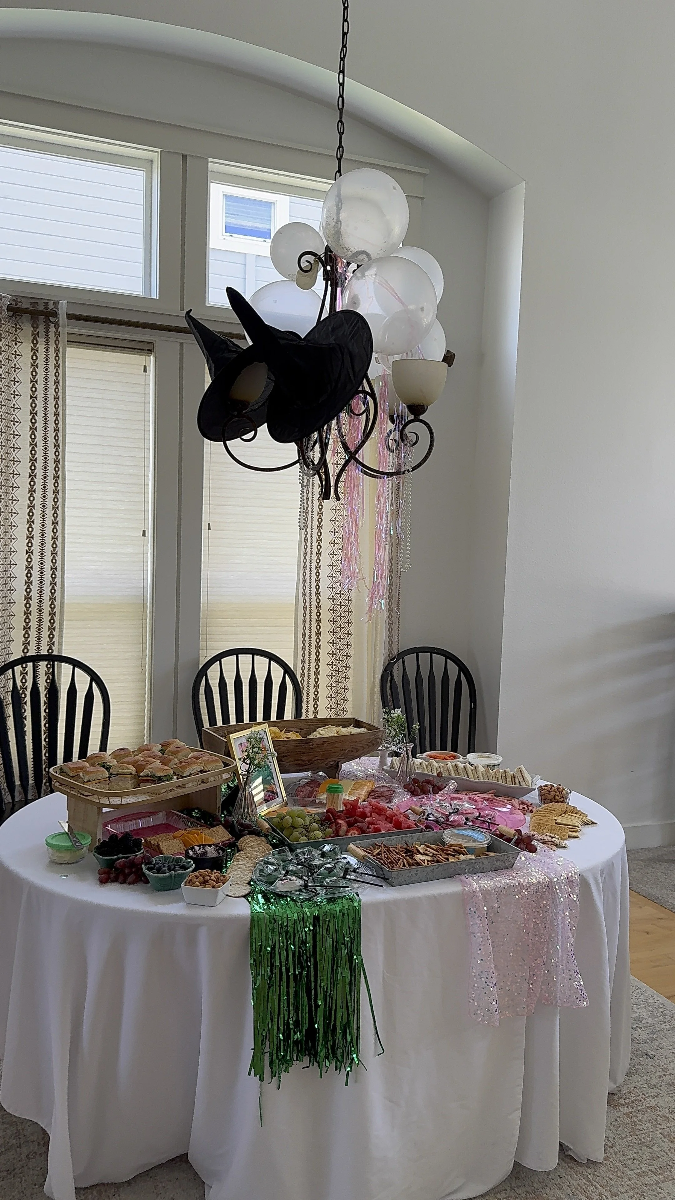 A table set with various snacks and fruits, decorated with pink and green streamers, under a chandelier with balloons and a witch hat.