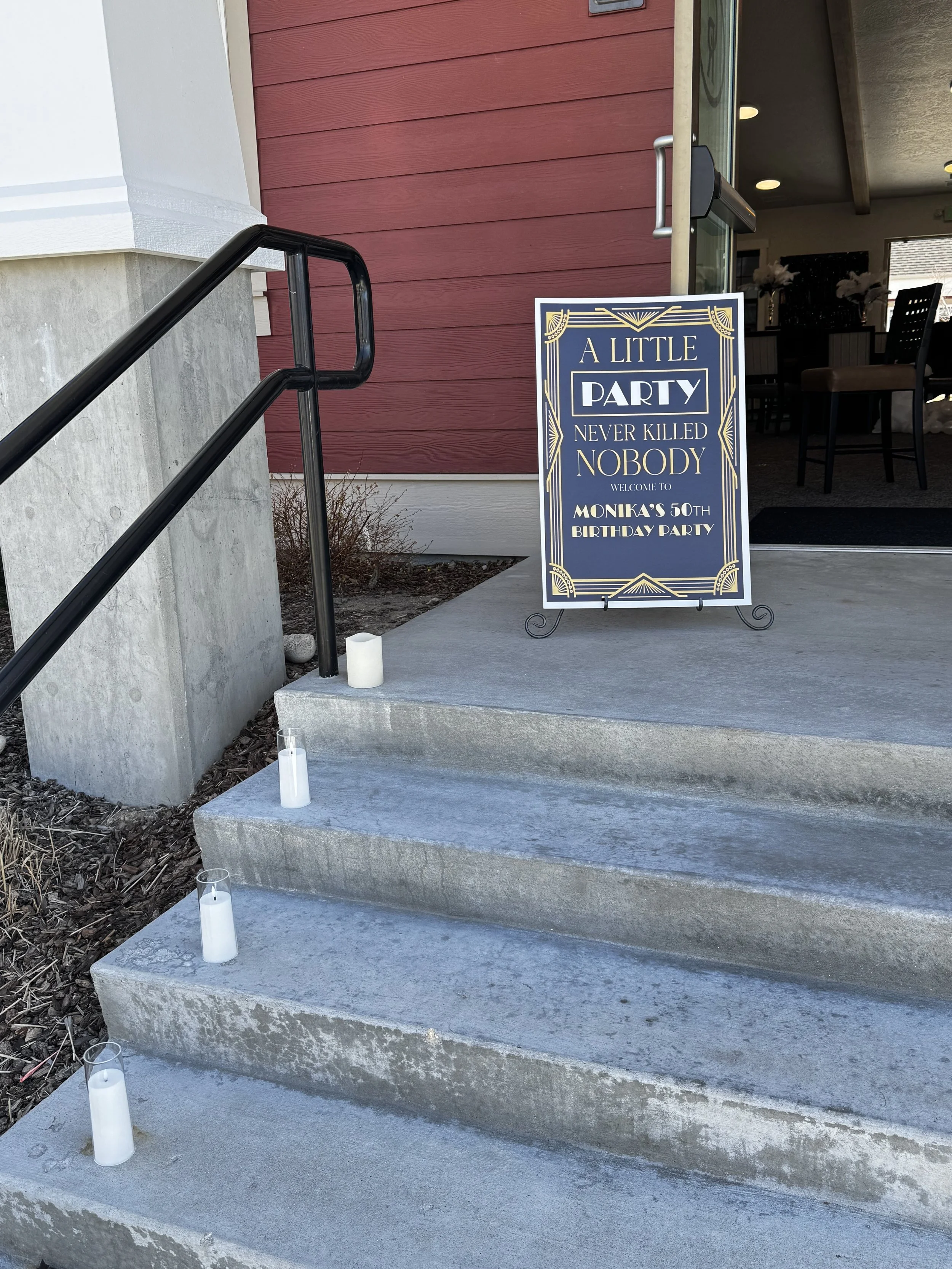A sign outside a building reads 'A LITTLE PARTY NEVER KILLED NOBODY' and announces 'WELCOME TO MONIKA'S 50TH BIRTHDAY PARTY'. The entrance has concrete steps with white candles in glass holders on each step, leading into a room with dark furniture an